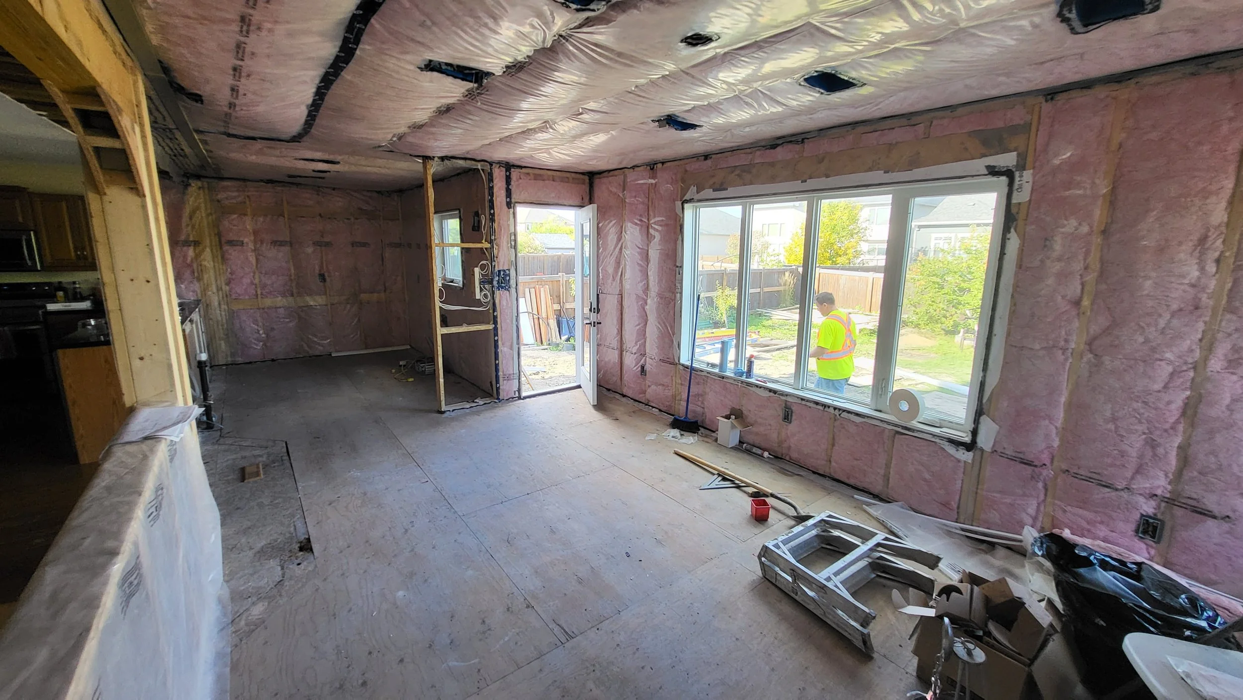 Interior view of a house under construction, showing pink insulation on the walls and ceiling, with open window and door, construction worker outside in a yellow safety vest, various construction materials and tools scattered on the floor.