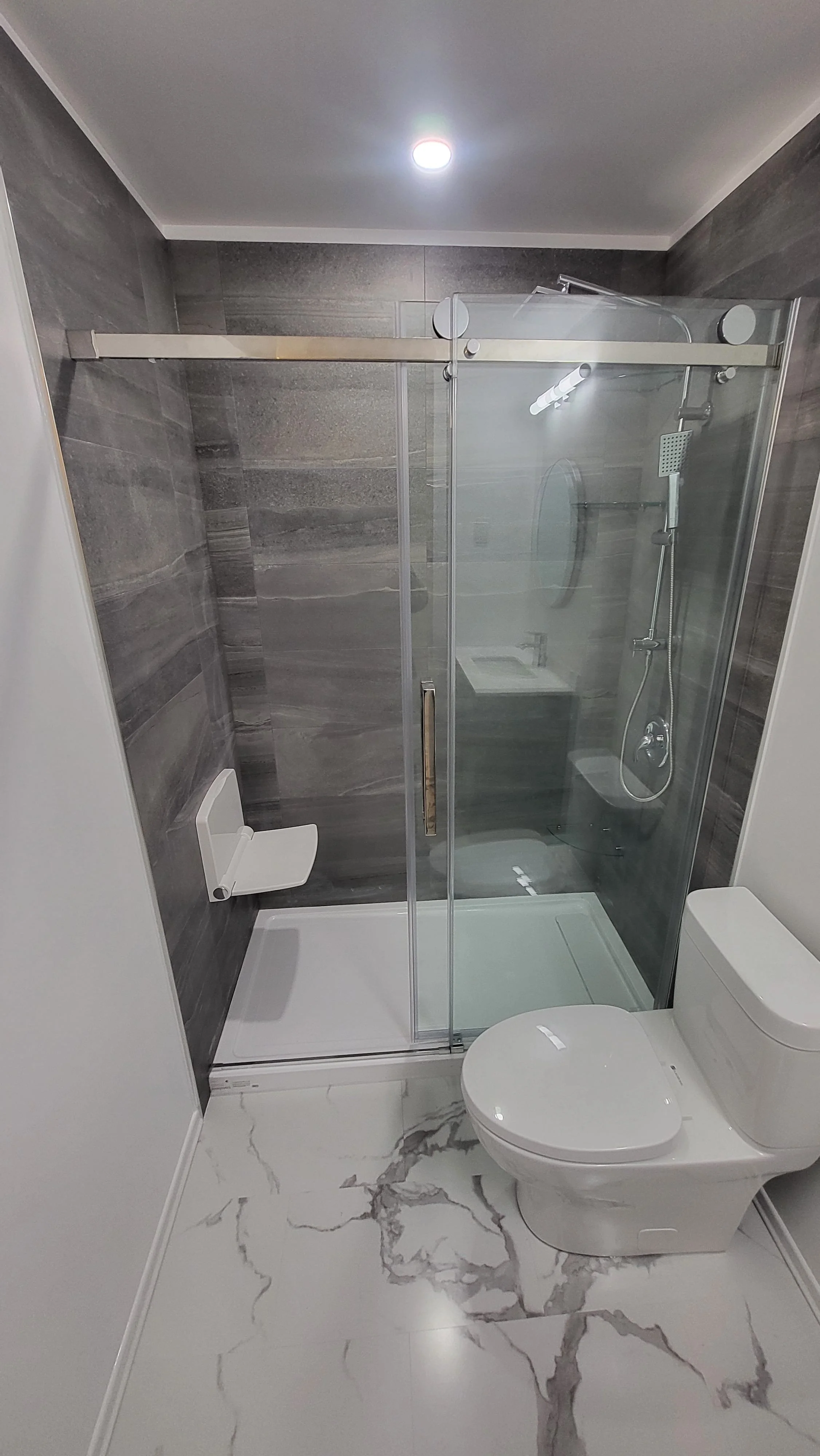 Modern bathroom with white toilet, marble floor, and glass shower stall with gray tiled walls. Bathroom contractor, tile installer, curb-less shower, glass shower doors