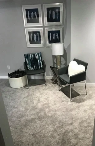Living room corner with two black chairs, a side table with a white lamp, a fluffy white pillow, a round white storage bin, and four framed abstract art pieces hanging on the wall.