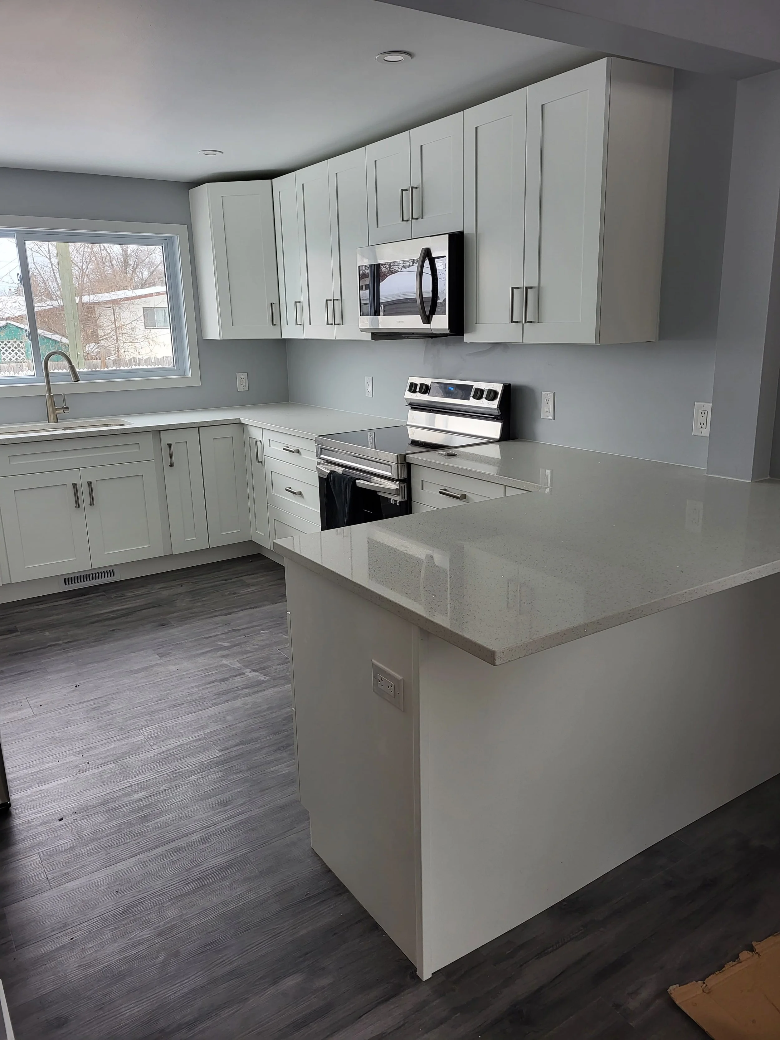 Modern kitchen with white cabinets, stainless steel appliances, a black oven, a microwave above, gray walls, a large window, and dark wood flooring. kitchen contractor, cabinet installer, appliance installer, drywall contractor, contractor.