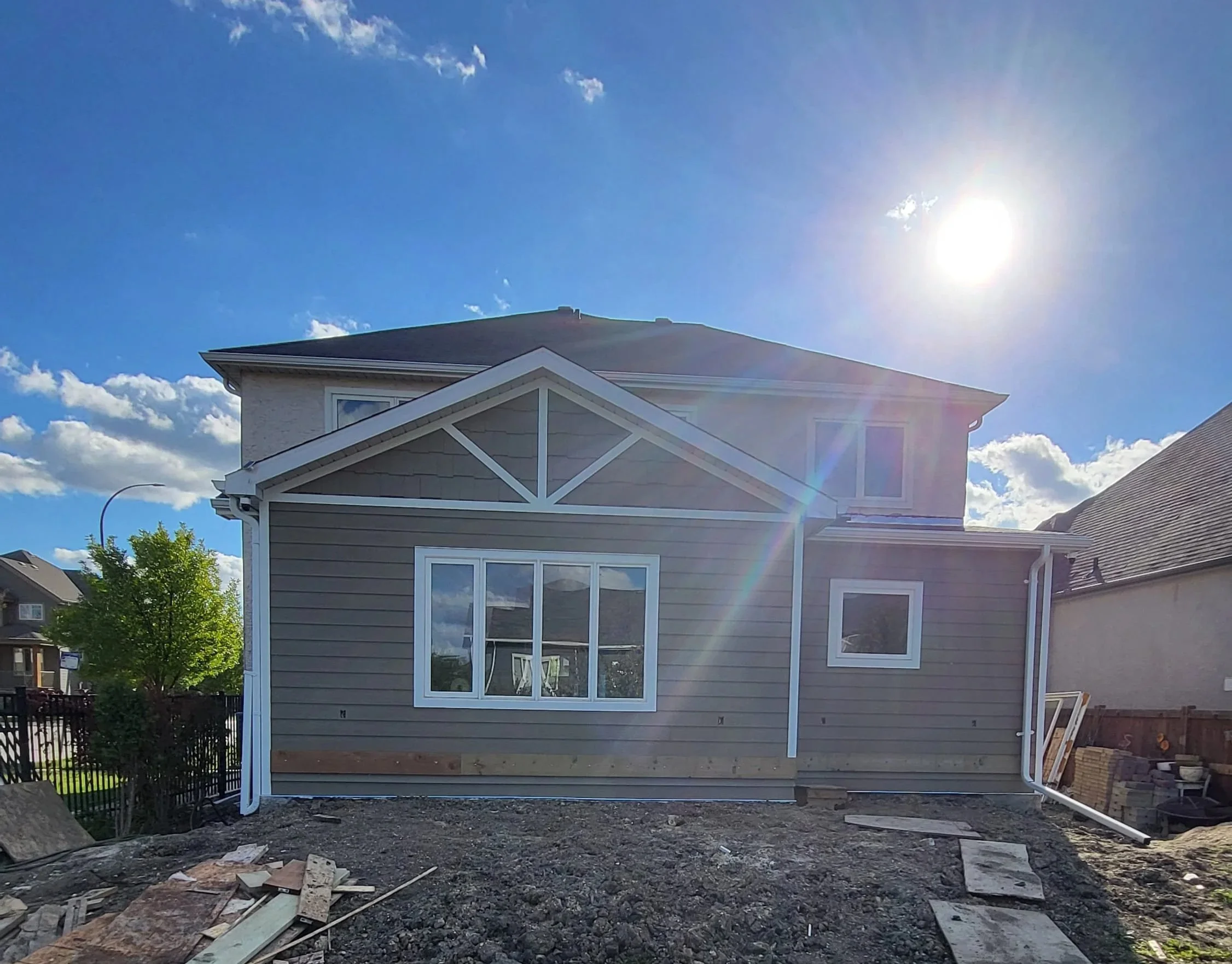 Rear view of a two-story house under construction with gray siding, white trim, and multiple windows, sun shining brightly in clear blue sky with some clouds. Home addition contractor
