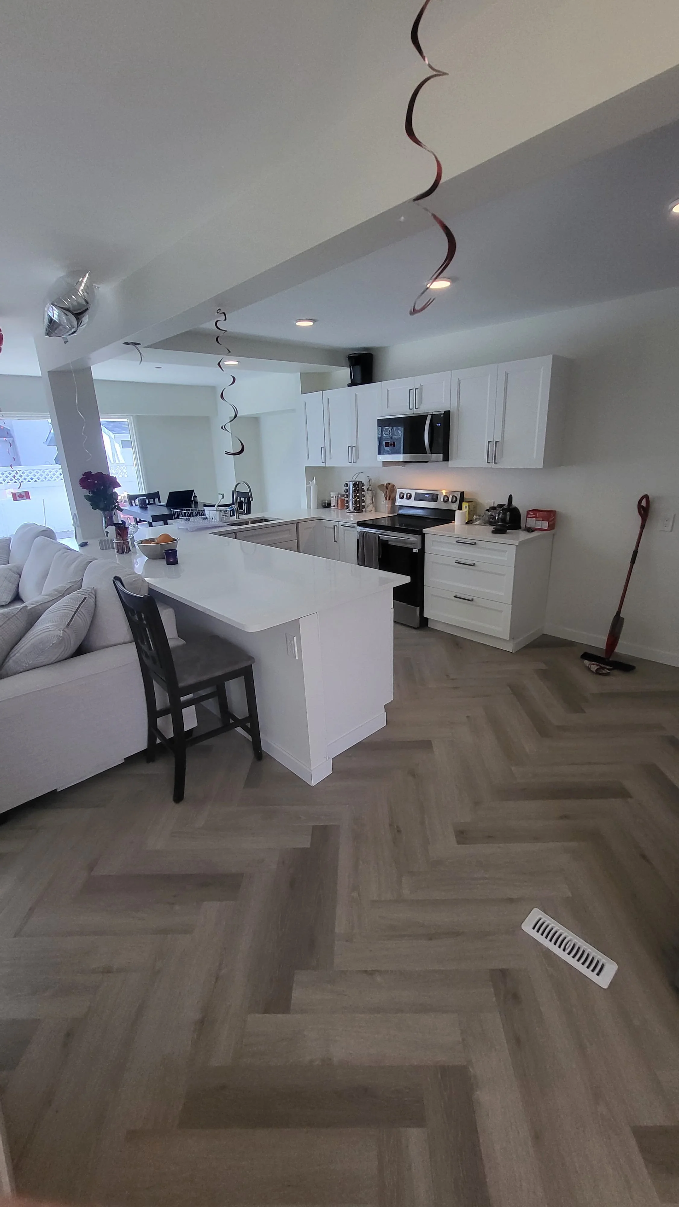 A modern kitchen with white cabinets, stainless steel appliances, and a large white island. There are decorations hanging from the ceiling, and a broom is leaning against the wall. The floor has a herringbone wood pattern. kitchen contractor, cabinet