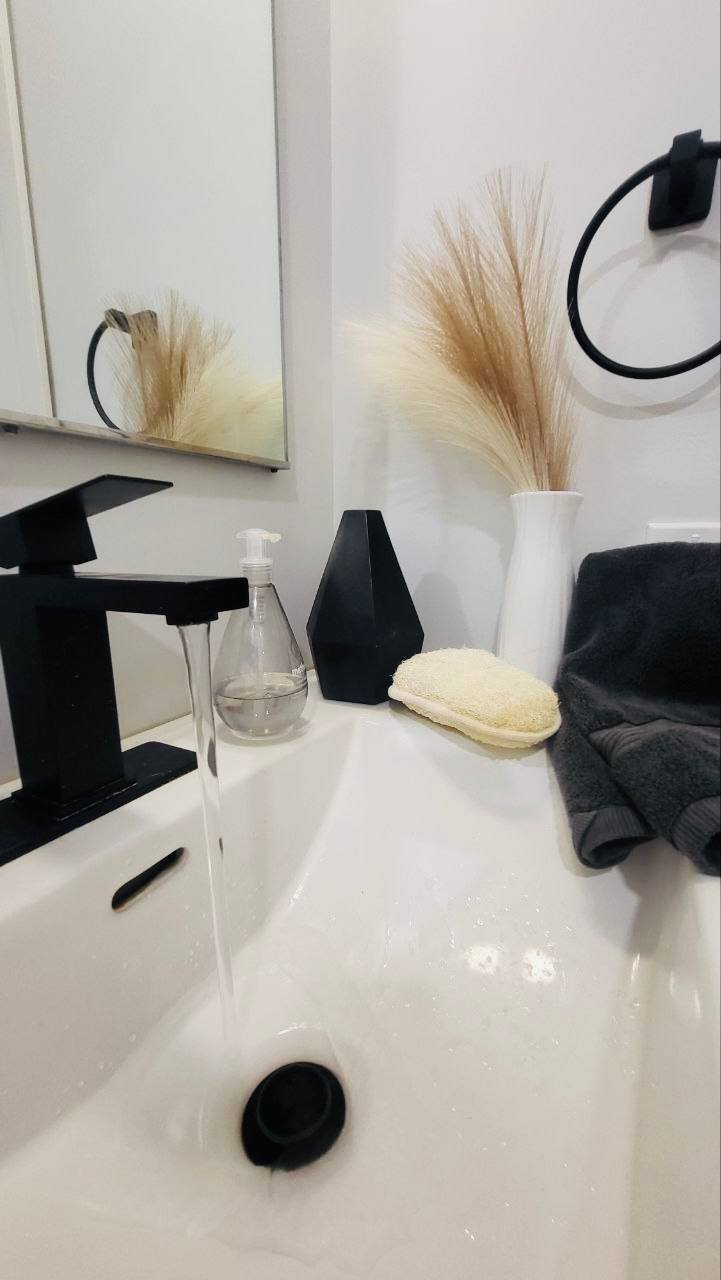 Bathroom sink with a black faucet, a clear soap dispenser, a black vase, a white vase with pampas grass, a folded towel, and a dark cloth on the right.