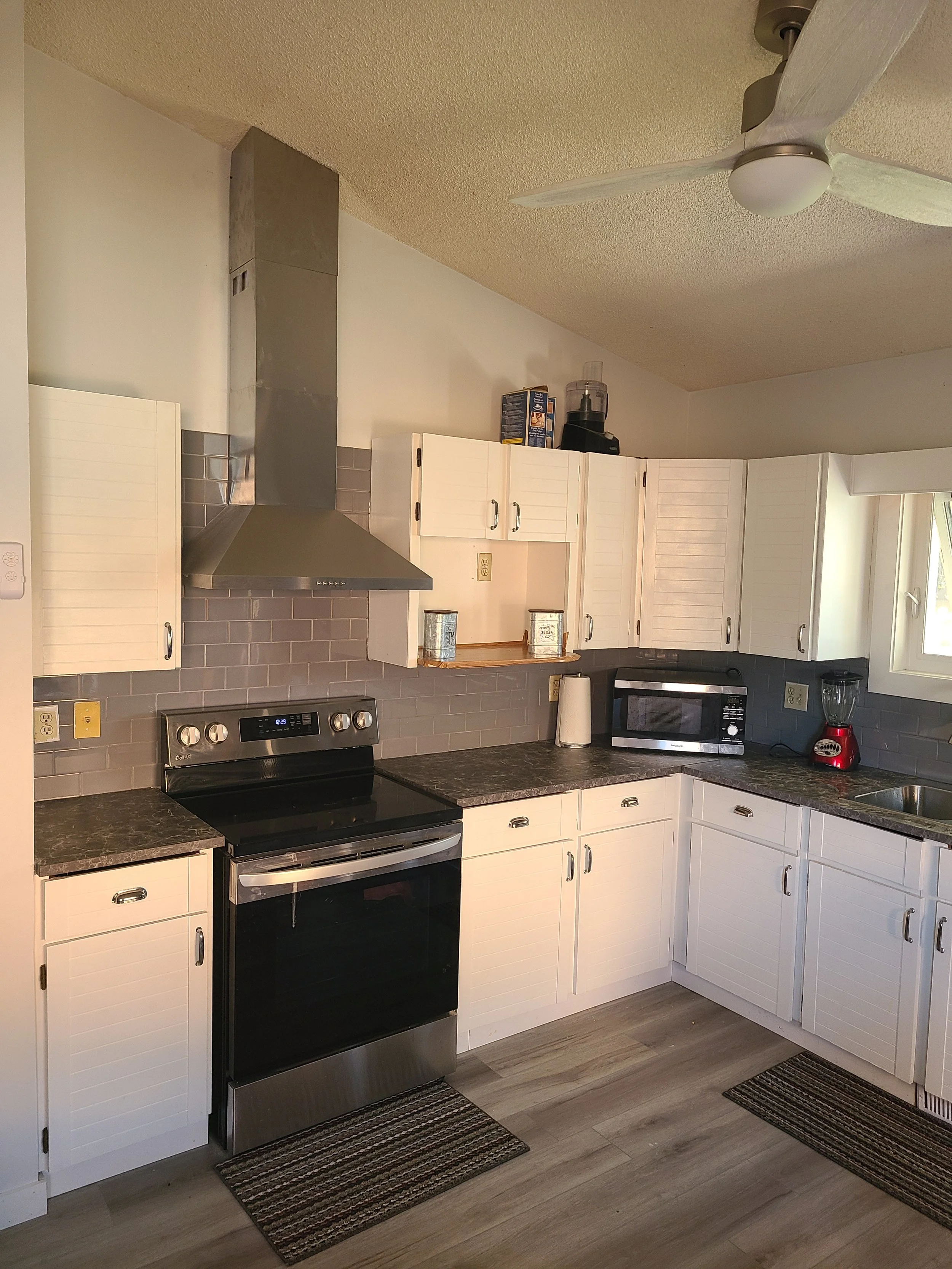 Kitchen with white cabinets, stainless steel stove, black countertop, gray tile backsplash, microwave, blender, paper towel holder, and window above the sink. Kitchen remodel.