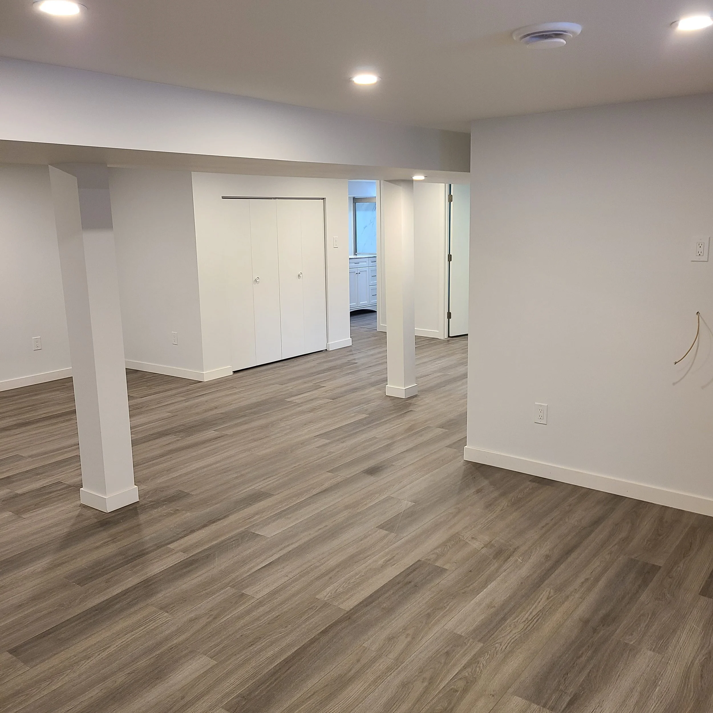 Empty room with wood-look flooring, white walls, recessed lighting, and a small kitchen at the back with white cabinetry.  Basement Renovation, hardwood floors, construction, drywall contractor, painter, framer