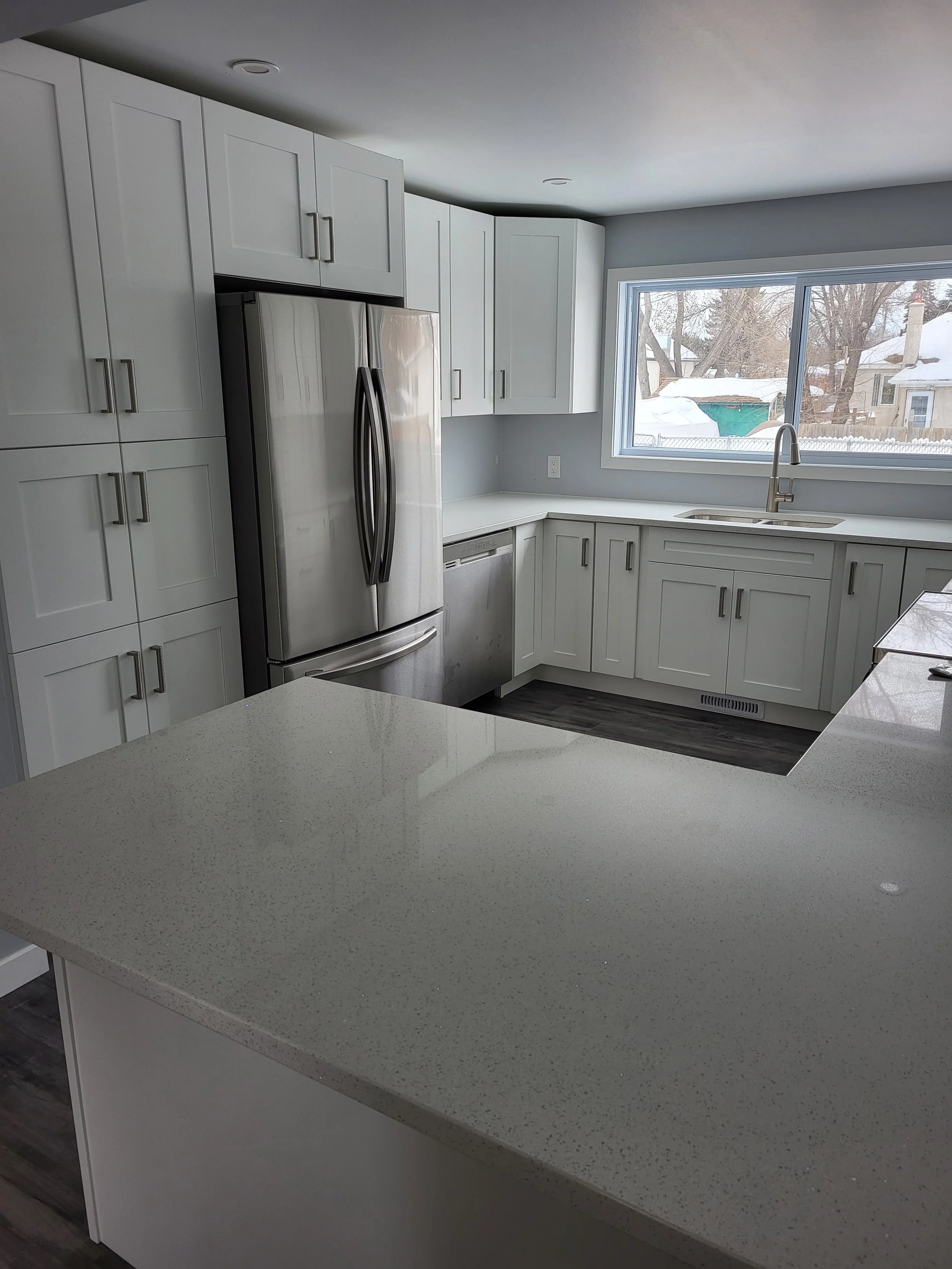 Modern white kitchen with stainless steel refrigerator, granite countertops, and a large window showing a snowy backyard. kitchen contractor, cabinet installer, appliance installer, drywall contractor, contractor.