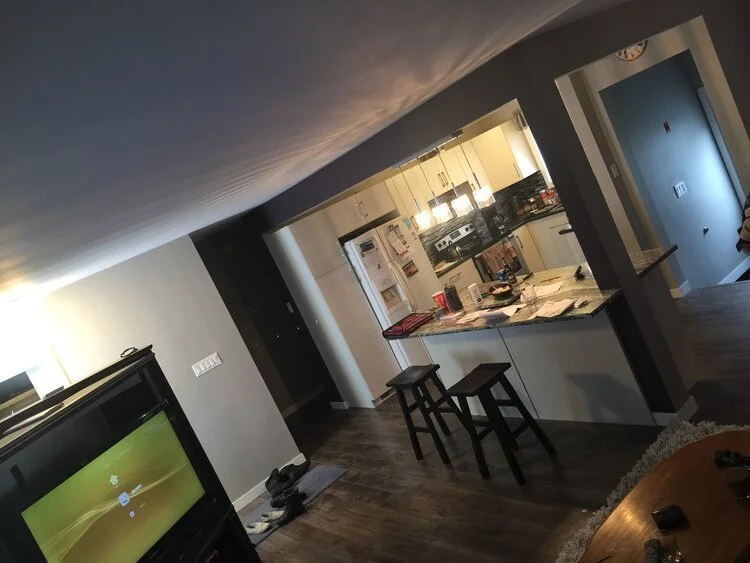 A kitchen and living room area with a television on the left, a small entryway in the back, and bar stools at a kitchen counter. The counter has various items, including papers and a soda can. The kitchen has white cabinets, a white refrigerator, and black countertops. The room has wood flooring and gray walls.