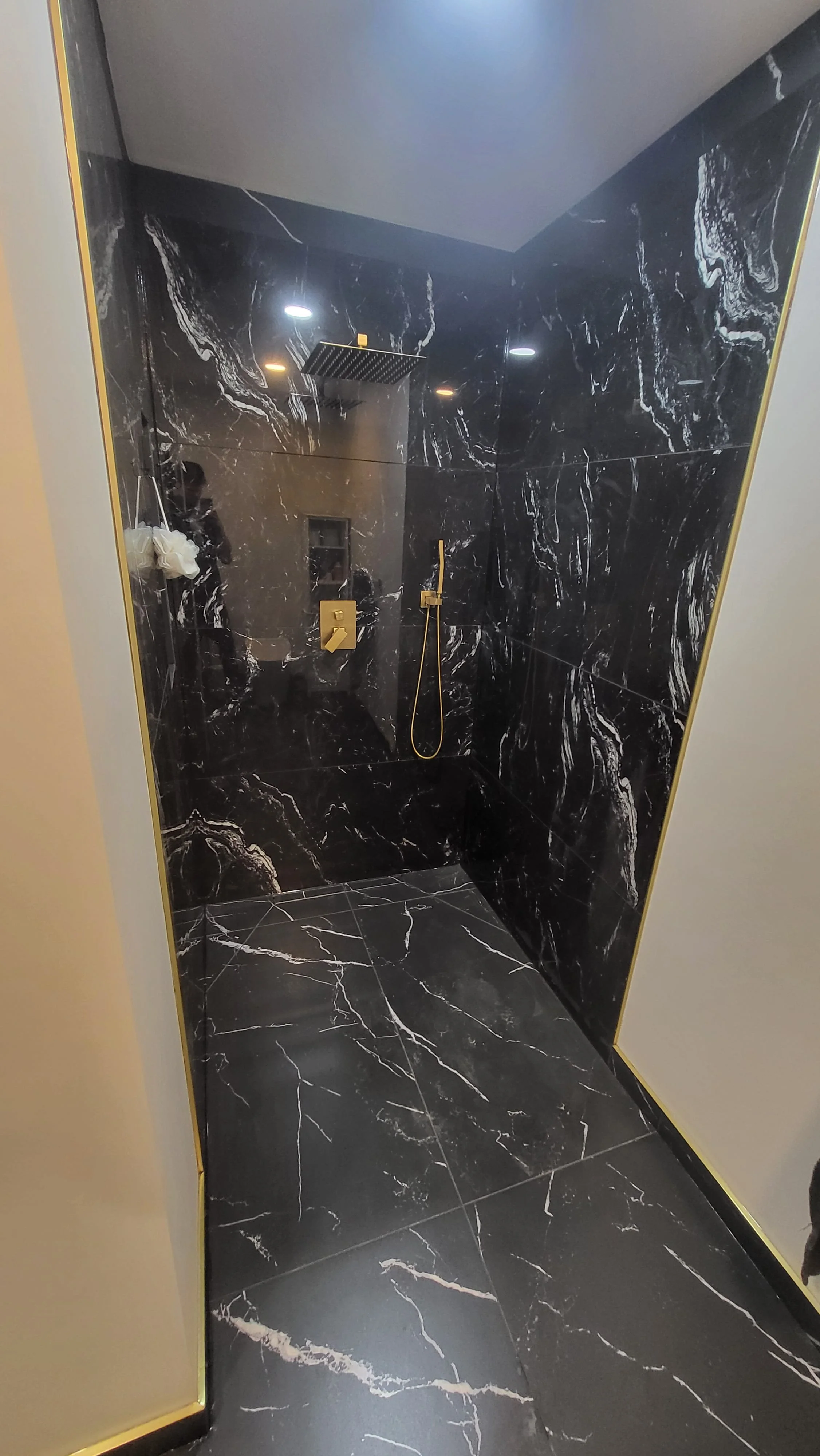 A modern shower with black marble tiles and gold fixtures, including a showerhead, handle, and handheld shower, with black marble flooring and a wall with a built-in niche for toiletries. Bathroom contractor, tile installer, curb-less shower, 
