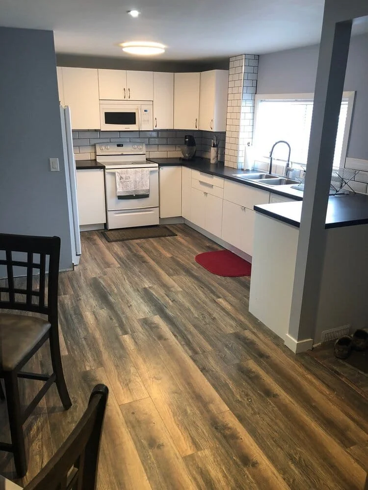 A kitchen with white cabinets, black countertops, a stove, microwave, sink by a window, and a wooden floor. There's a small red mat in front of the sink. kitchen contractor, cabinet installer, appliance installer, drywall contractor, contractor.