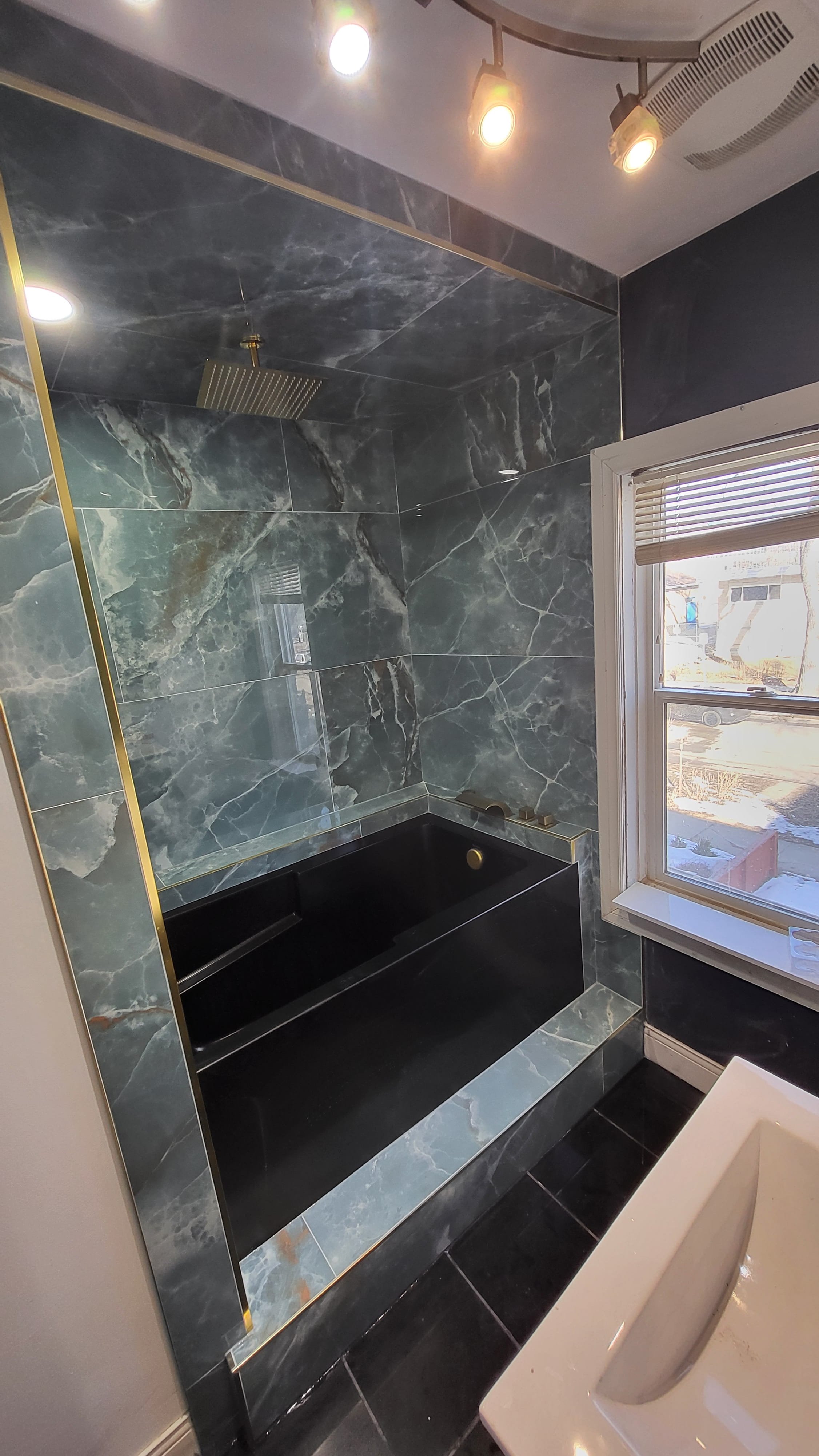 Bathroom with dark green marble walls and a black bathtub, a window with blinds, and a ceiling light fixture. Bathroom contractor, tile installer, curb-less shower, glass shower doors