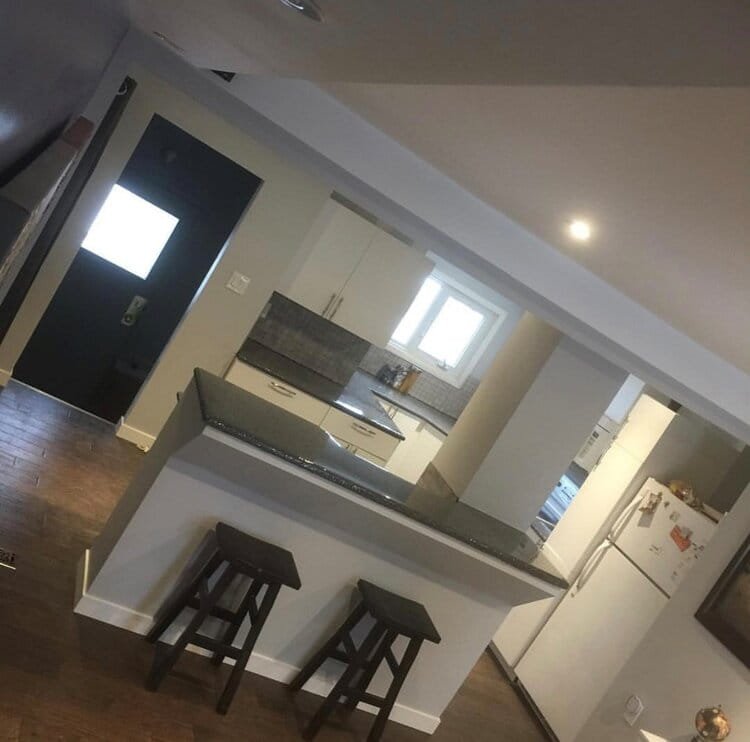 A modern kitchen with a breakfast bar, two wooden stools, white cabinets, a gray countertop, a window, a refrigerator, and a black door with a window. kitchen contractor, cabinet installer, appliance installer, drywall contractor, contractor.