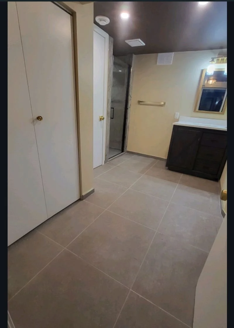 A bathroom with beige walls, a shower with glass door, a dark-colored vanity with a mirror, and large grey floor tiles. Bathroom contractor, tile installer, curb-less shower, glass shower doors