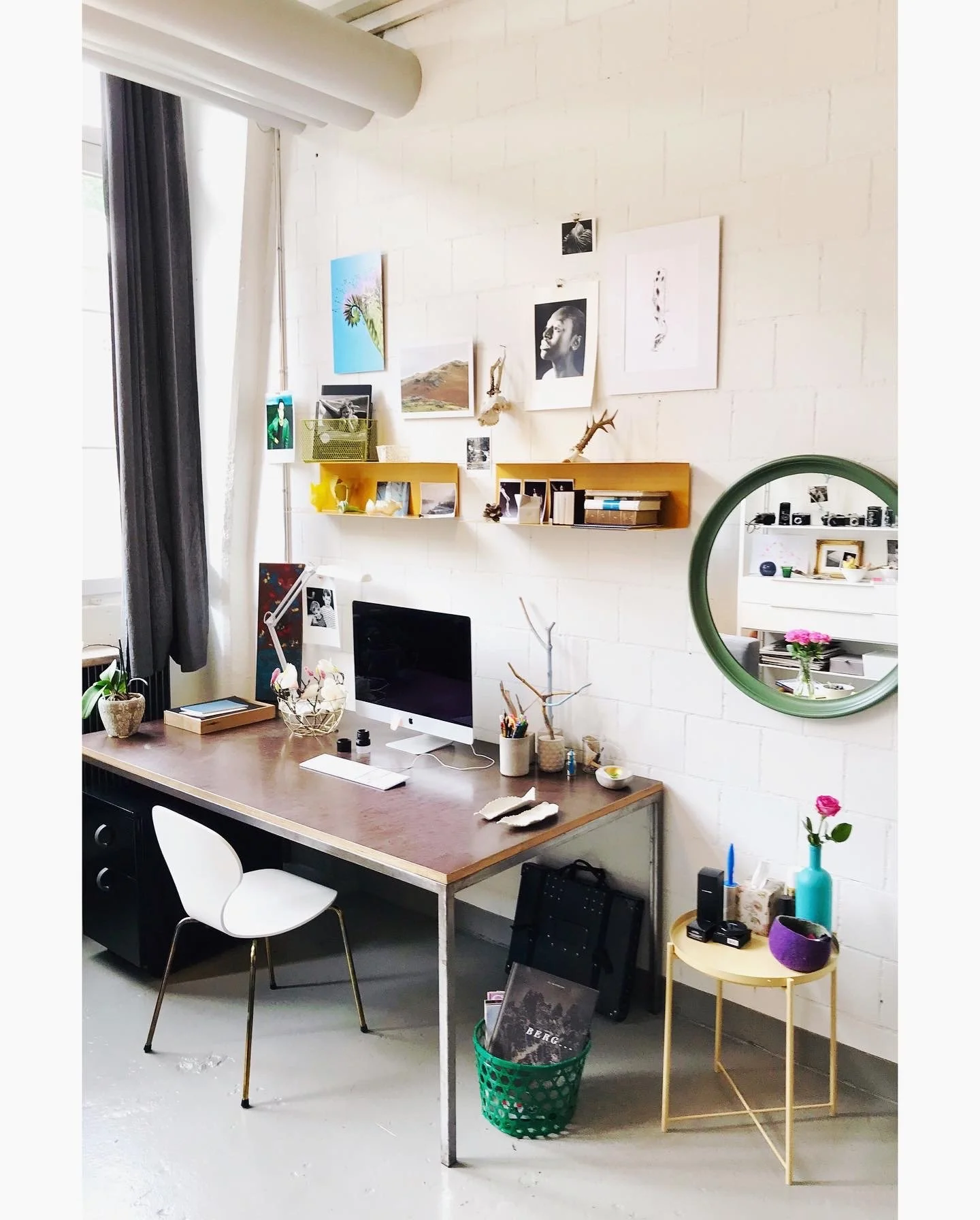 Ein heller Arbeitsbereich mit einem großen Tisch, einem Desktop-Computer, einer weißen Lampe und mehreren Pflanzen, vor einer Wand mit Kunstwerken und dekorativen Gegenständen.