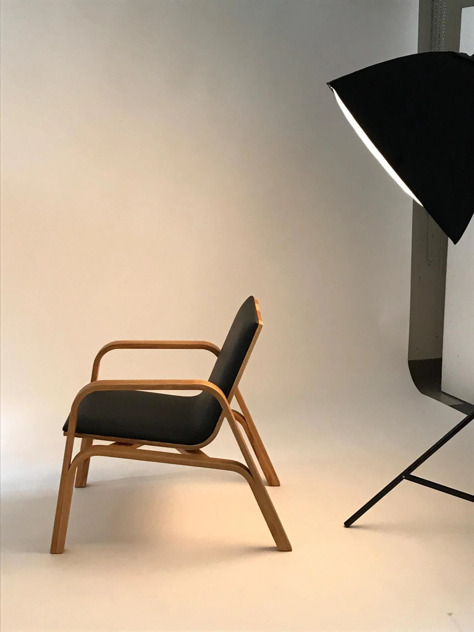 Ein einzelner Stuhl mit Holzrahmen und schwarzer Sitzfläche, neben einem Studio- oder Fotolicht auf einem weißen Hintergrund.