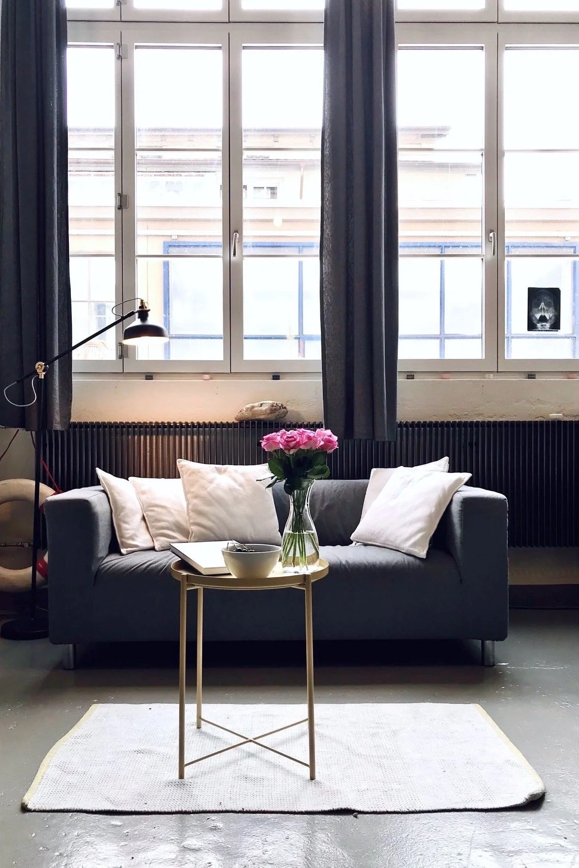 Gemütliches Wohnzimmer mit grauem Sofa, weißen Kissen, einem kleinen Beistelltisch mit Vase und Rosen, großen Fenstern mit dunklen Vorhängen, weißen Wänden und einem dunklen Heizkörper.