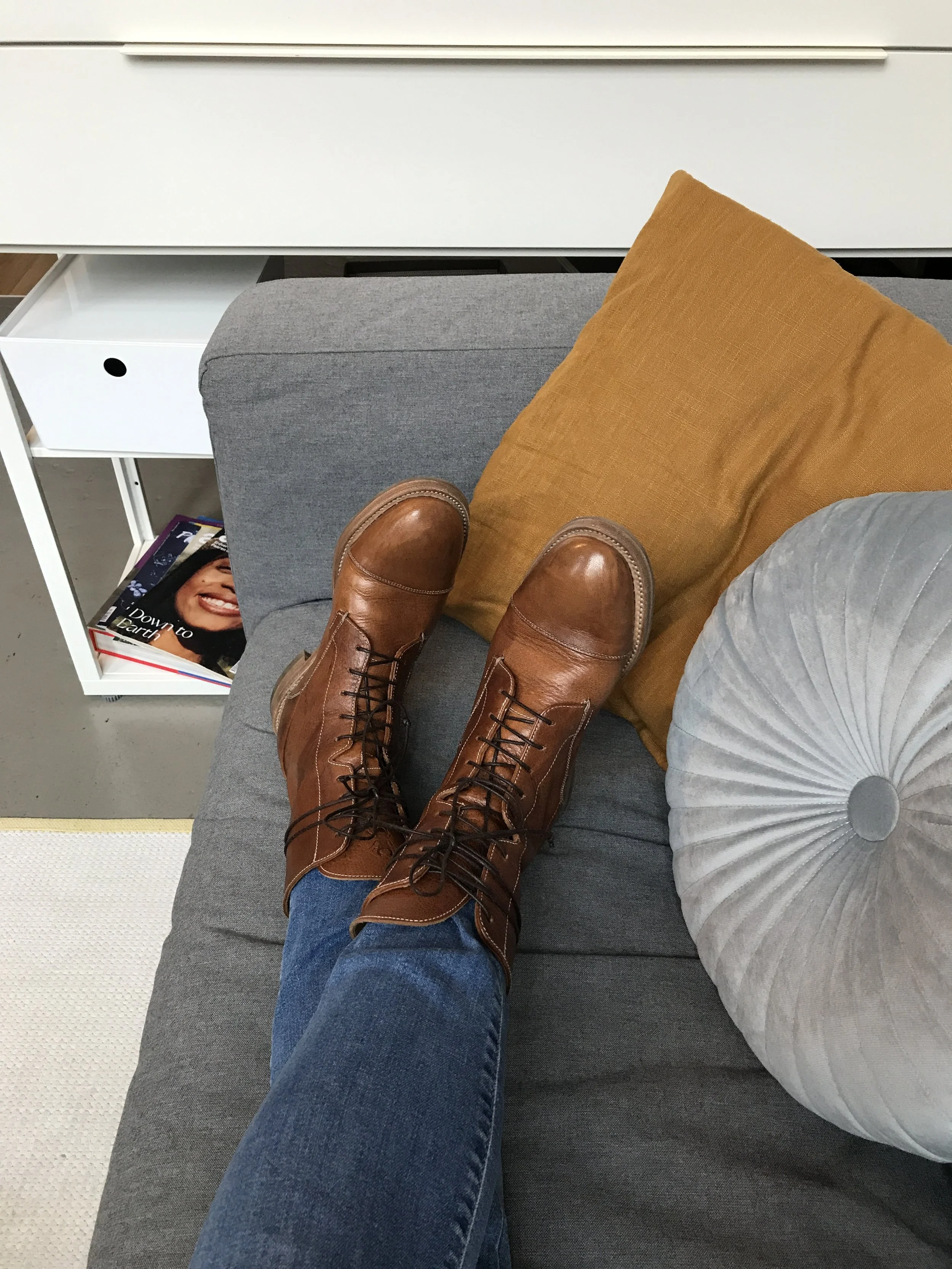 Perspektive auf die Beine einer Person, die braune Stiefel trägt, auf einem grauen Sofa sitzend, mit einem goldenen Kissen und einem hellgrauen Kissen und einem weißen Beistelltisch mit Büchern im Hintergrund.