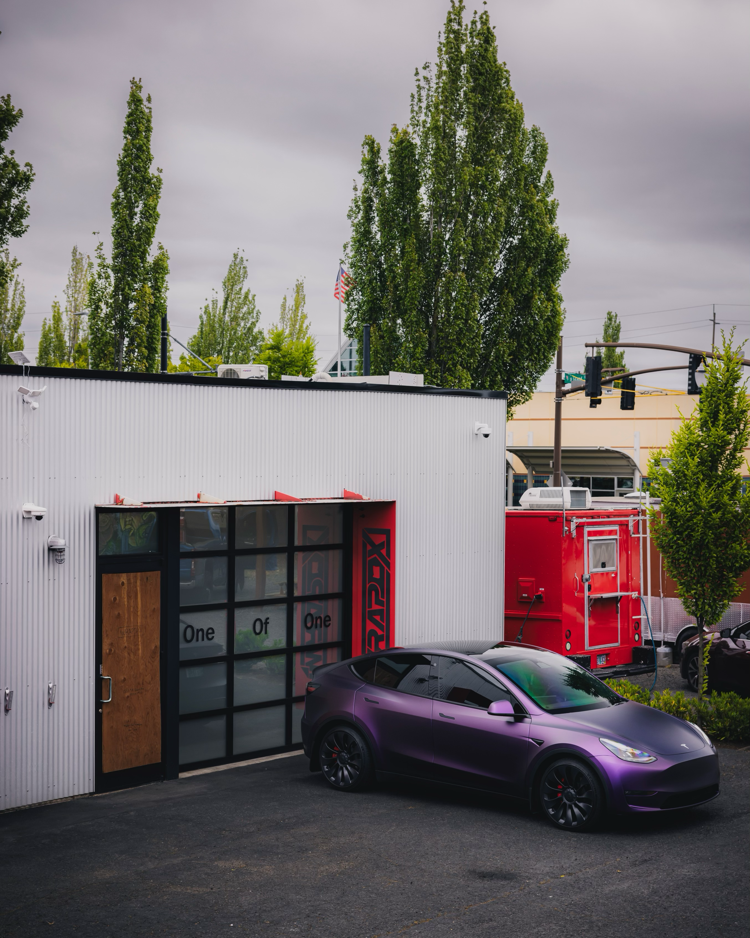A matte purple Tesla parked outside a modern building with a glass garage door, surrounded by green trees and overcast sky.