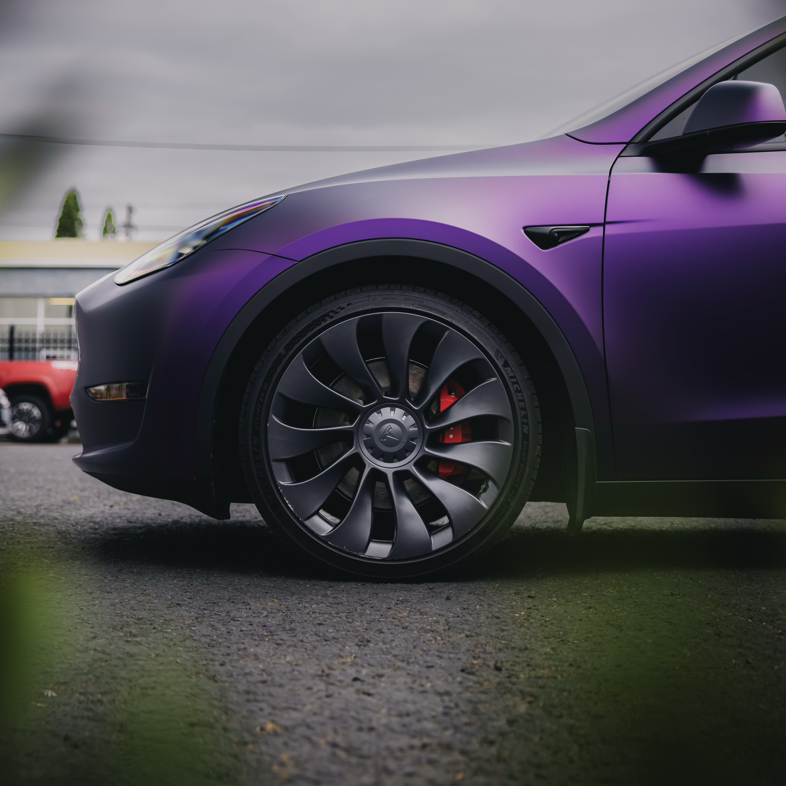 Side view of a purple car with a focus on the front wheel with dark colored alloy rims and red brake calipers, parked on an asphalt surface.