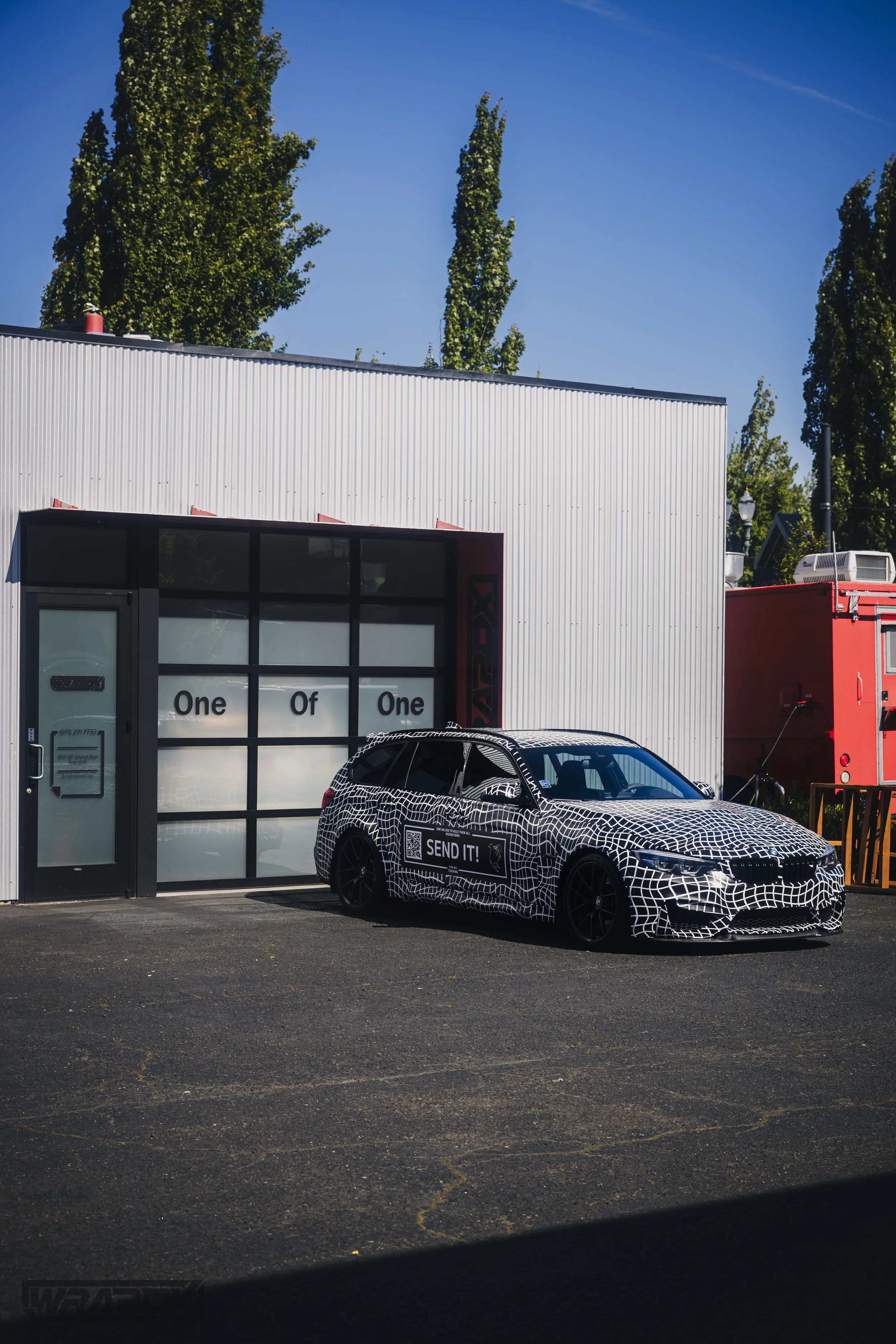A car with a black-and-white geometric wrap parked in front of a modern building, with the words 'One Of One' on the building's window. Trees and a red structure are visible in the background.