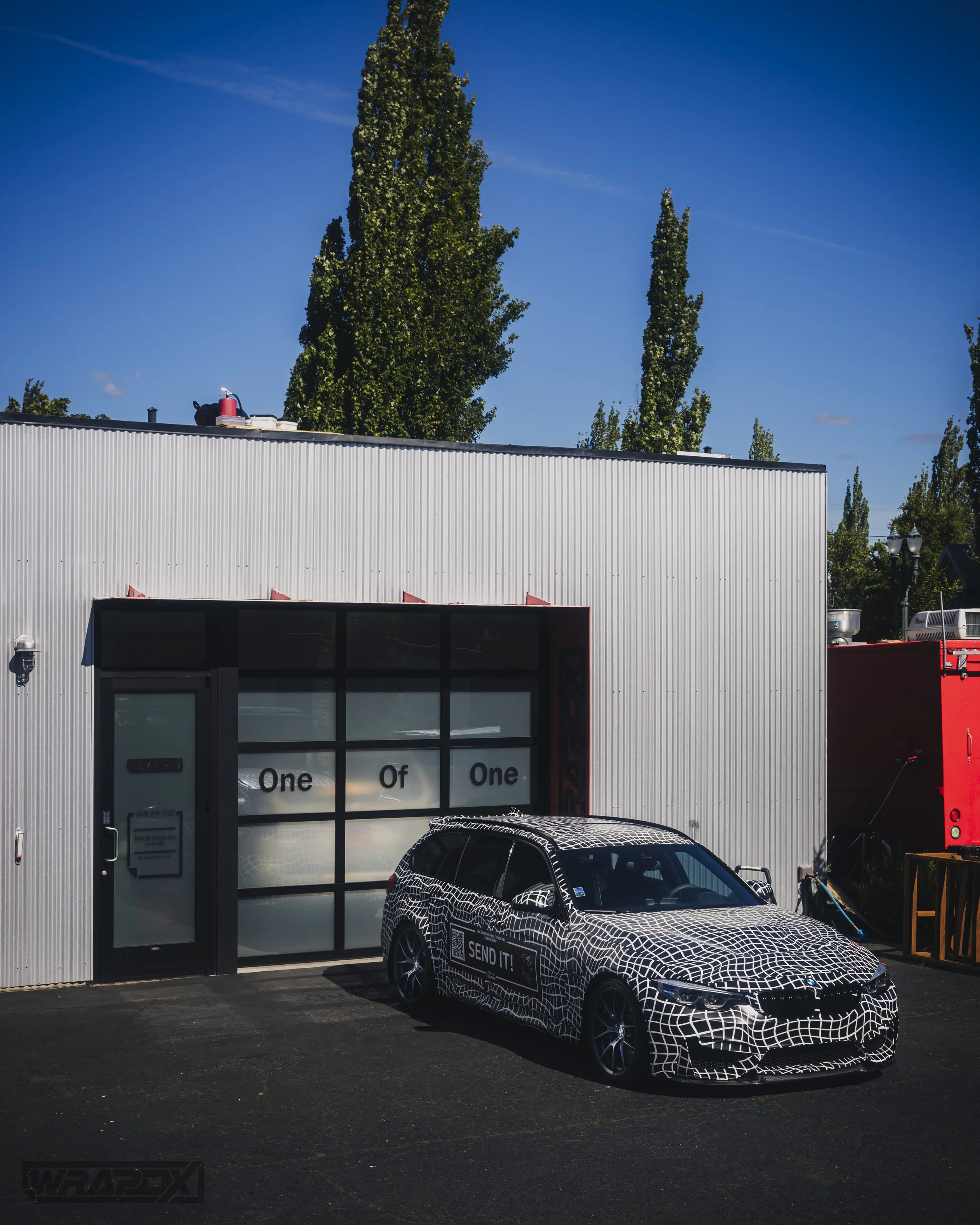 A car covered in a black and white geometric pattern, parked in front of a garage with "One of One" written on the door, next to a building with corrugated metal siding and trees in the background.