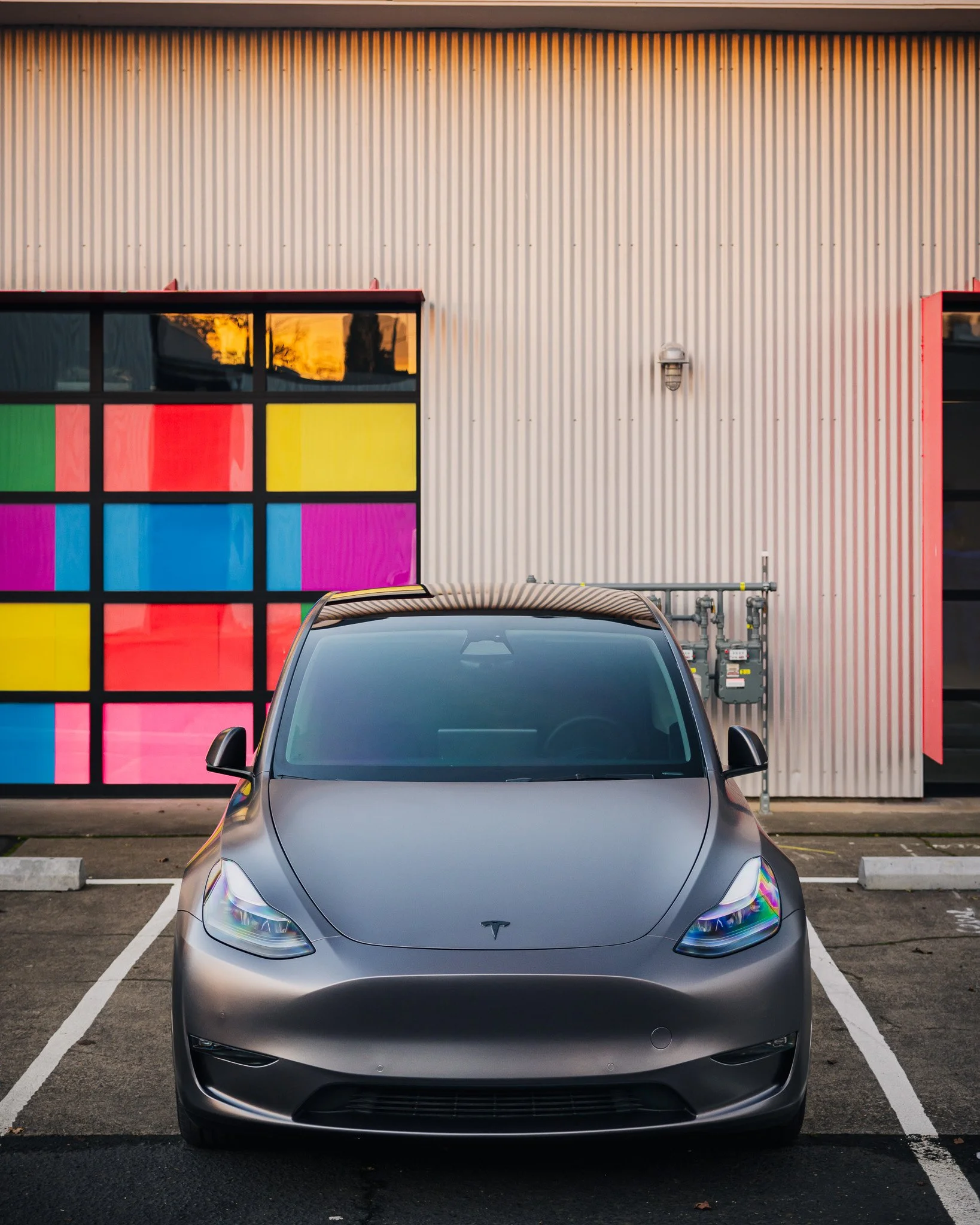 Front view of a gray electric car parked in front of a building with a colorful glass panel wall.