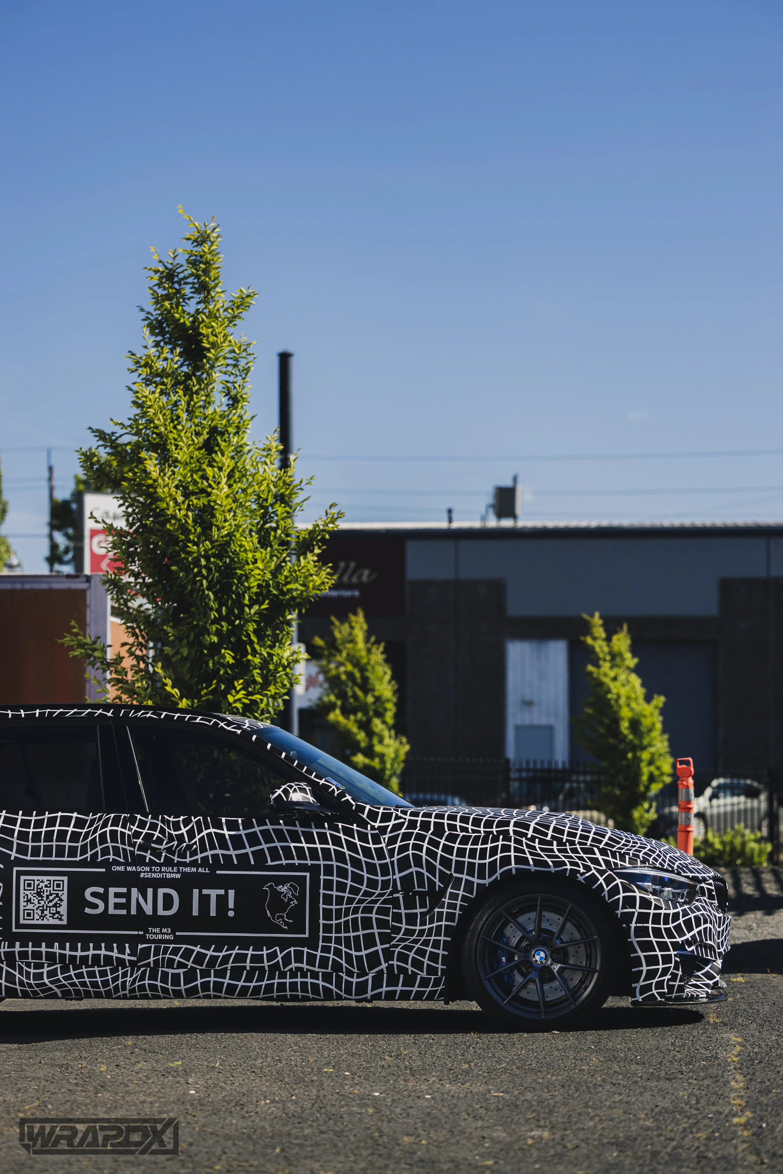 A black and white camouflaged car parked outdoors; featuring QR code and 'SEND IT!' text on the side. Surrounded by green trees and industrial buildings.