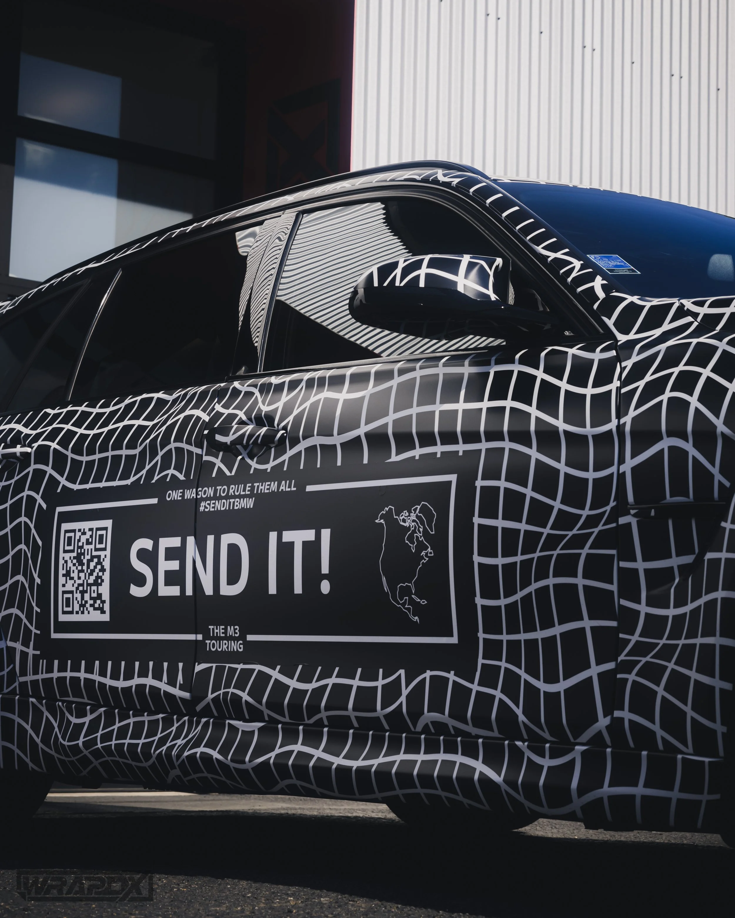Car with black and white camouflage design featuring "Send It!" text and QR code, side view.
