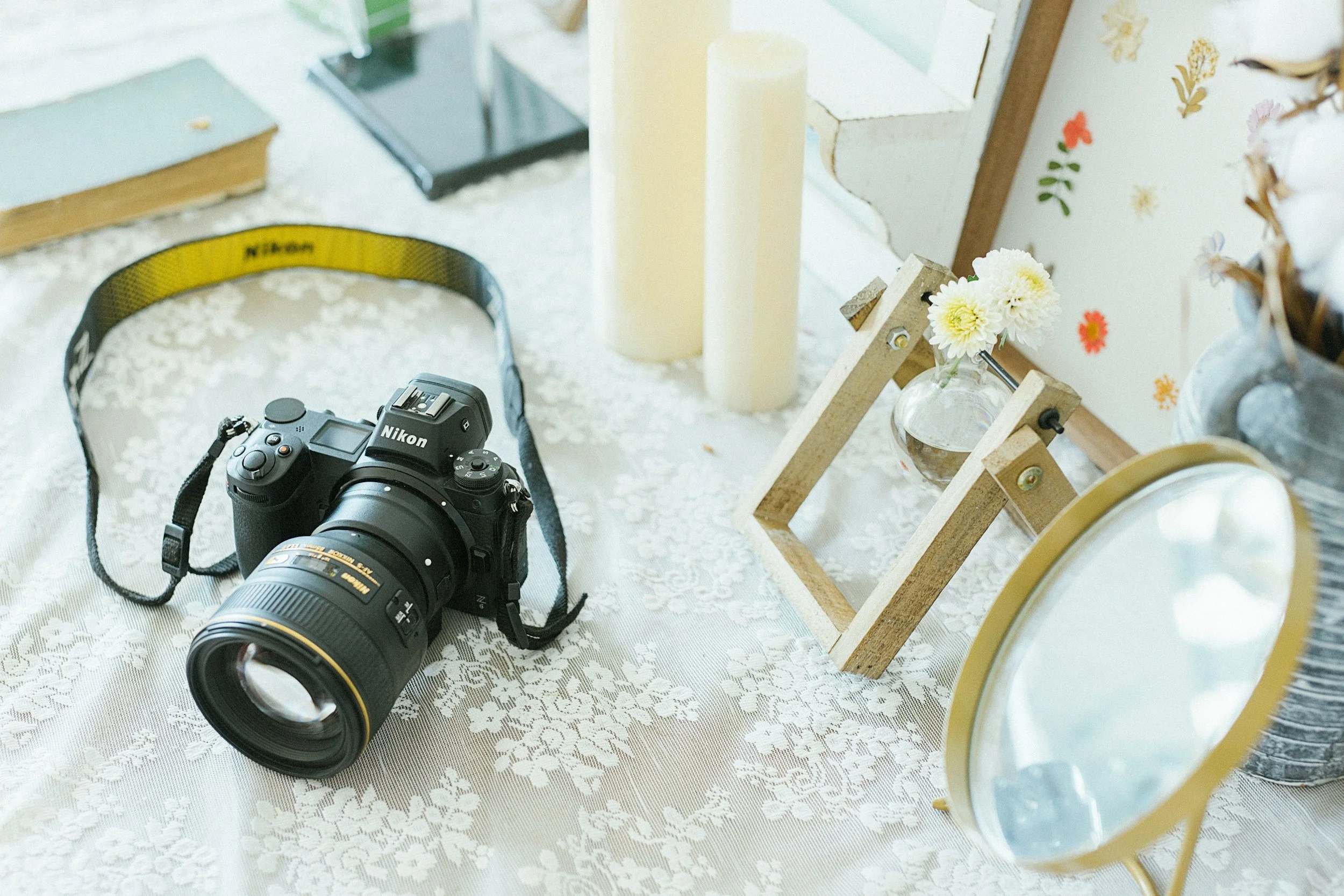 A Nikon DSLR camera with a zoom lens placed on a white lace tablecloth, alongside decorative items including candles, a small glass vase with white flowers in a wooden frame, a mirror, and a basket with more flowers.