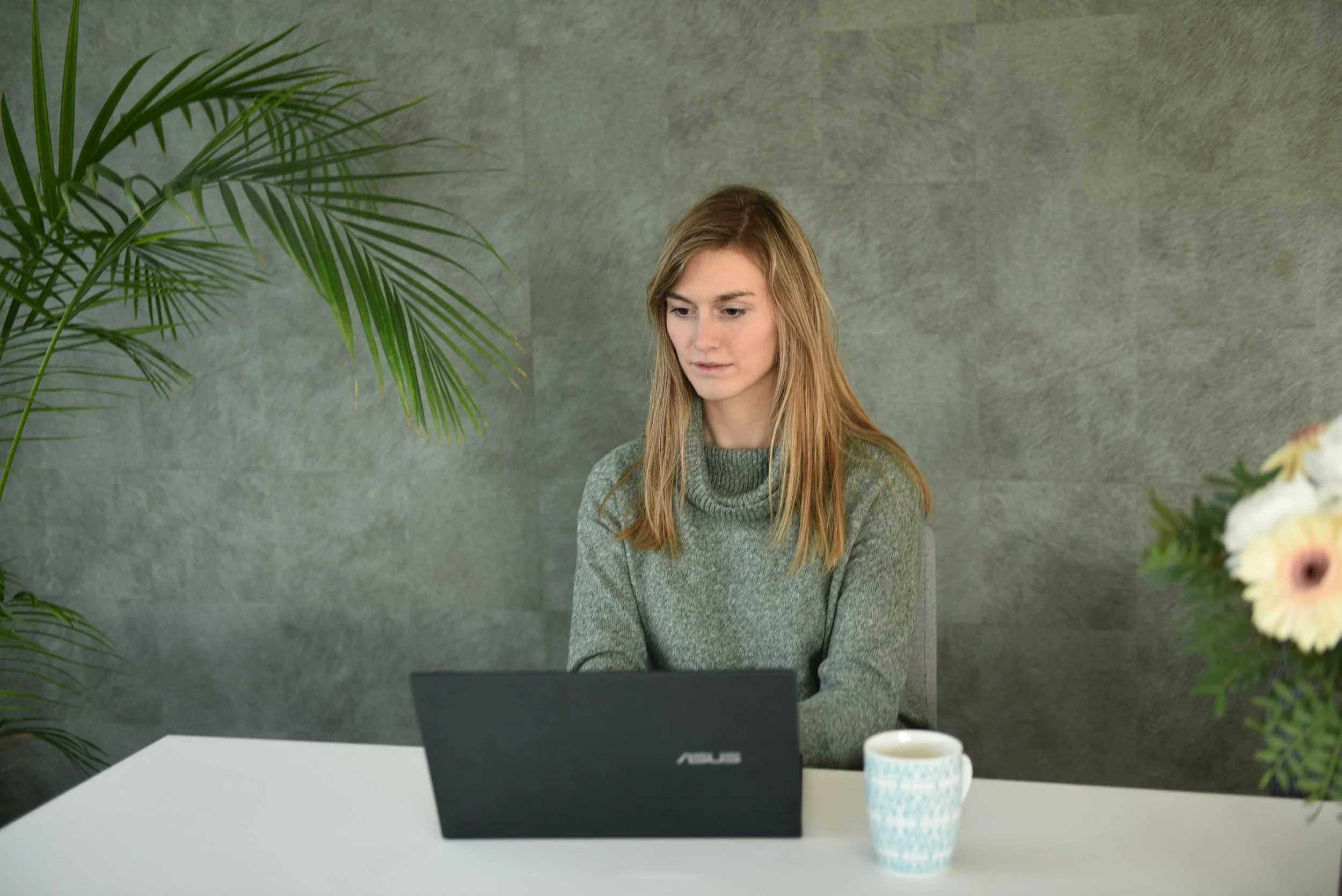 A woman sitting at a white table working on a black ASUS laptop, with a white mug and greenery in the background.