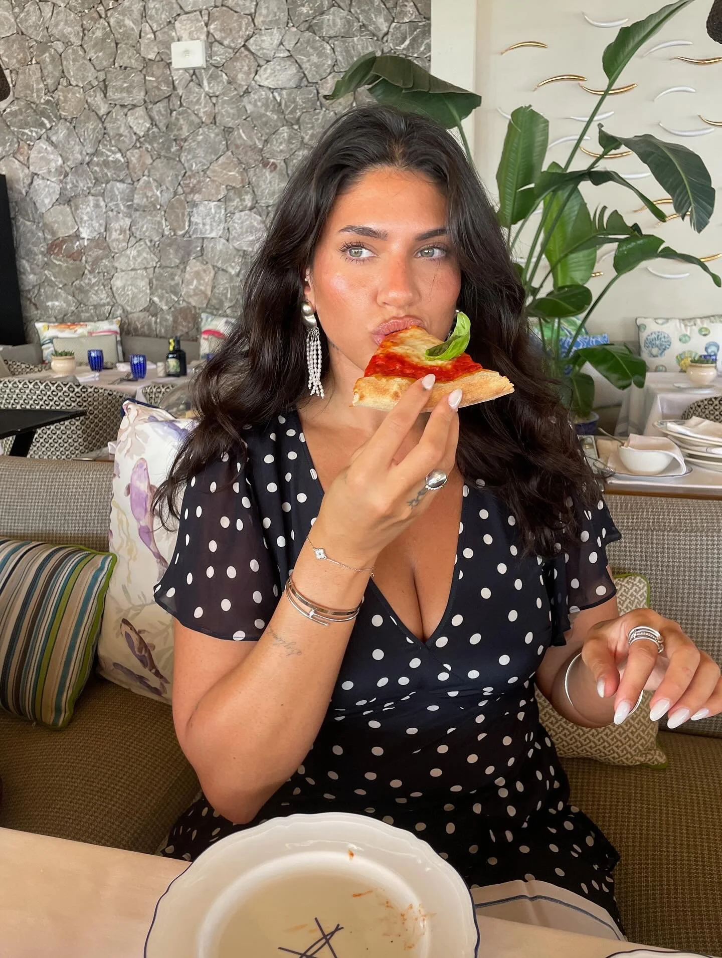 A woman with long dark hair, wearing a black dress with white polka dots, is holding a slice of pizza with tomato sauce, cheese, and basil. She is sitting in a restaurant with a stone textured wall, green plants, and decorated cushions.