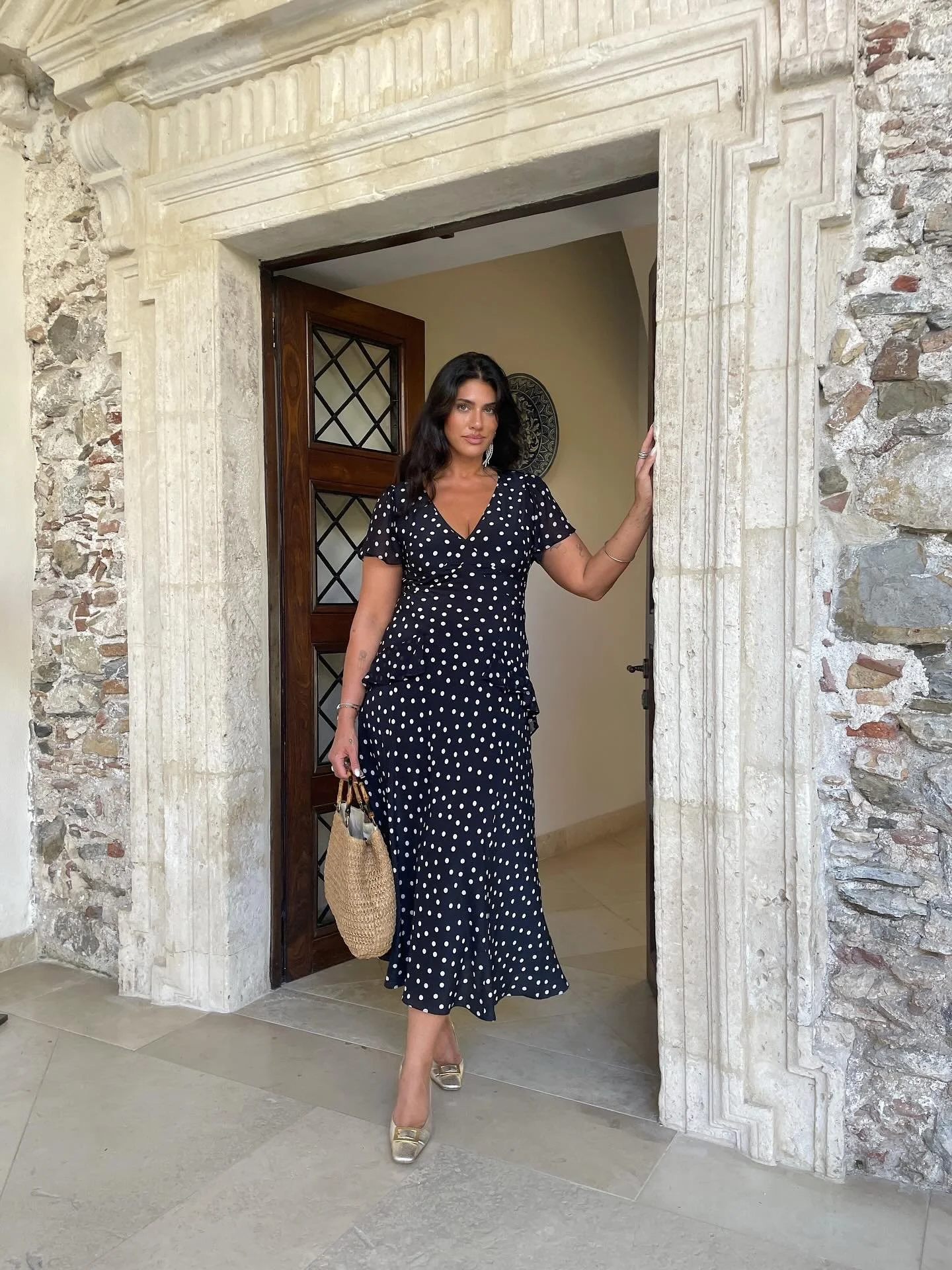 A woman in a navy blue polka dot dress stands in a doorway with a stone arch, holding a woven handbag and wearing gold shoes.