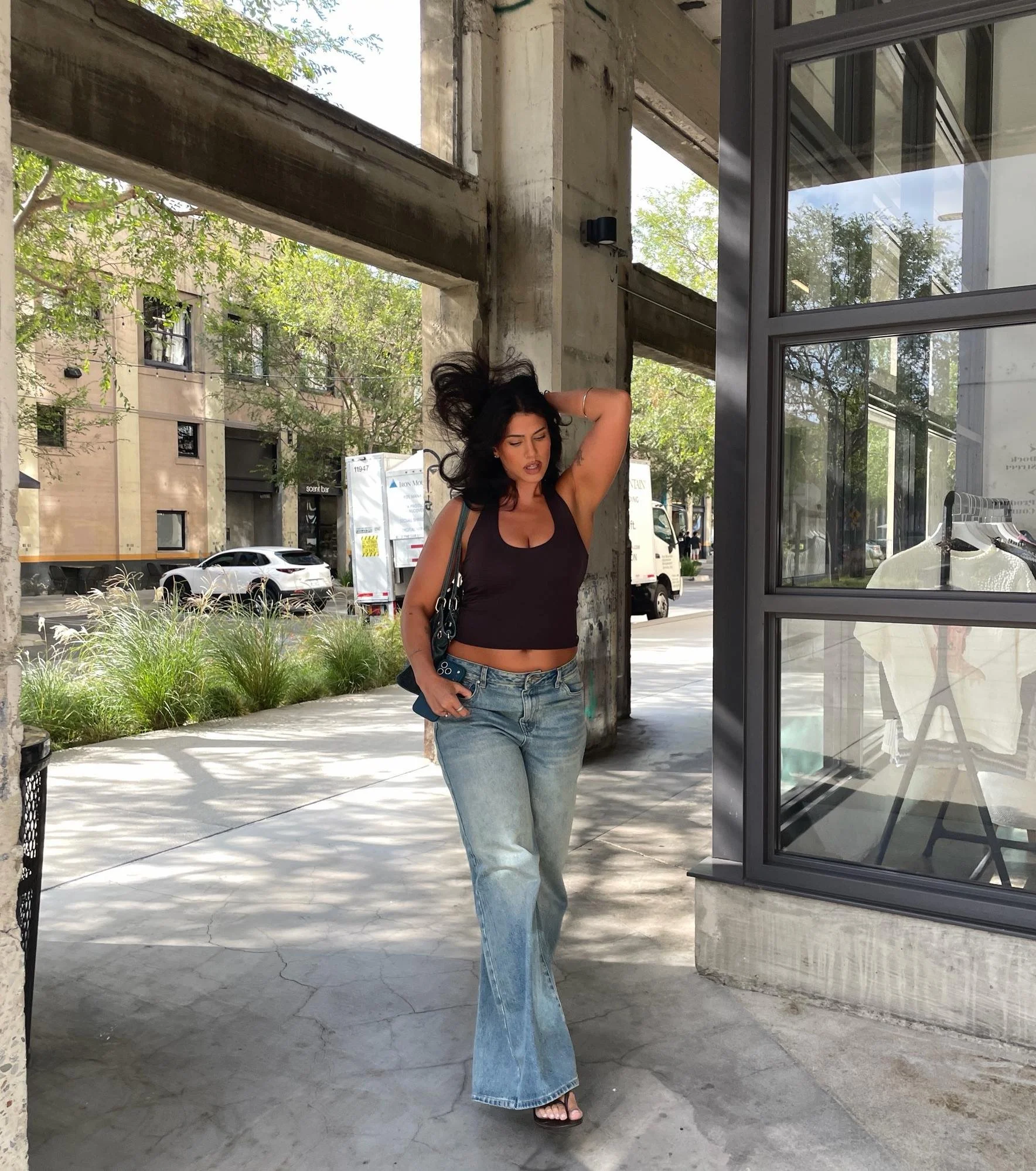 A woman standing outside near a building with large glass windows, holding her hair back with one hand and wearing a black crop top, blue jeans, and sandals. There are trees and parked cars in the background.