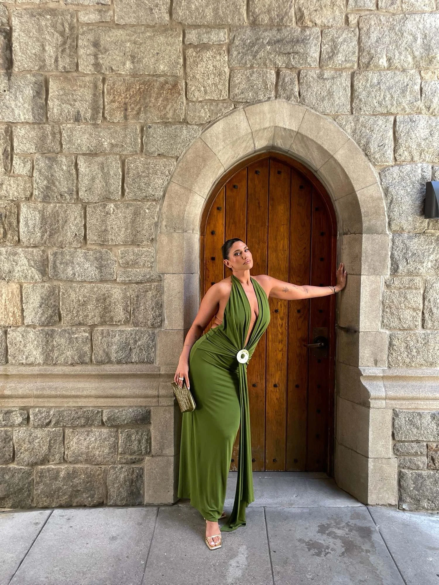A woman in a green dress standing in front of a stone wall and wooden door, posing with one hand on the wall and looking at the camera.