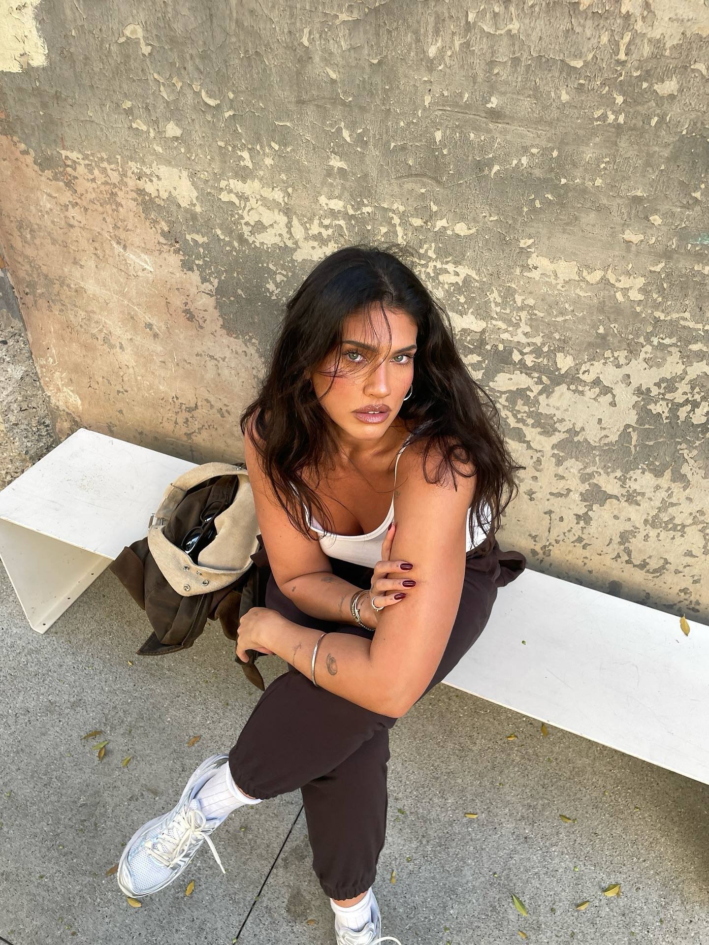 A woman with dark wavy hair sitting on a white bench against a weathered wall, wearing a white tank top, black pants, and white sneakers, with her arms crossed and looking at the camera.