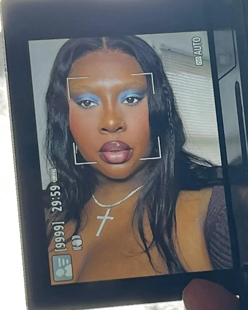 Photograph of a woman with dark hair and makeup, wearing a cross necklace, being taken on a camera or phone screen with various camera settings visible.