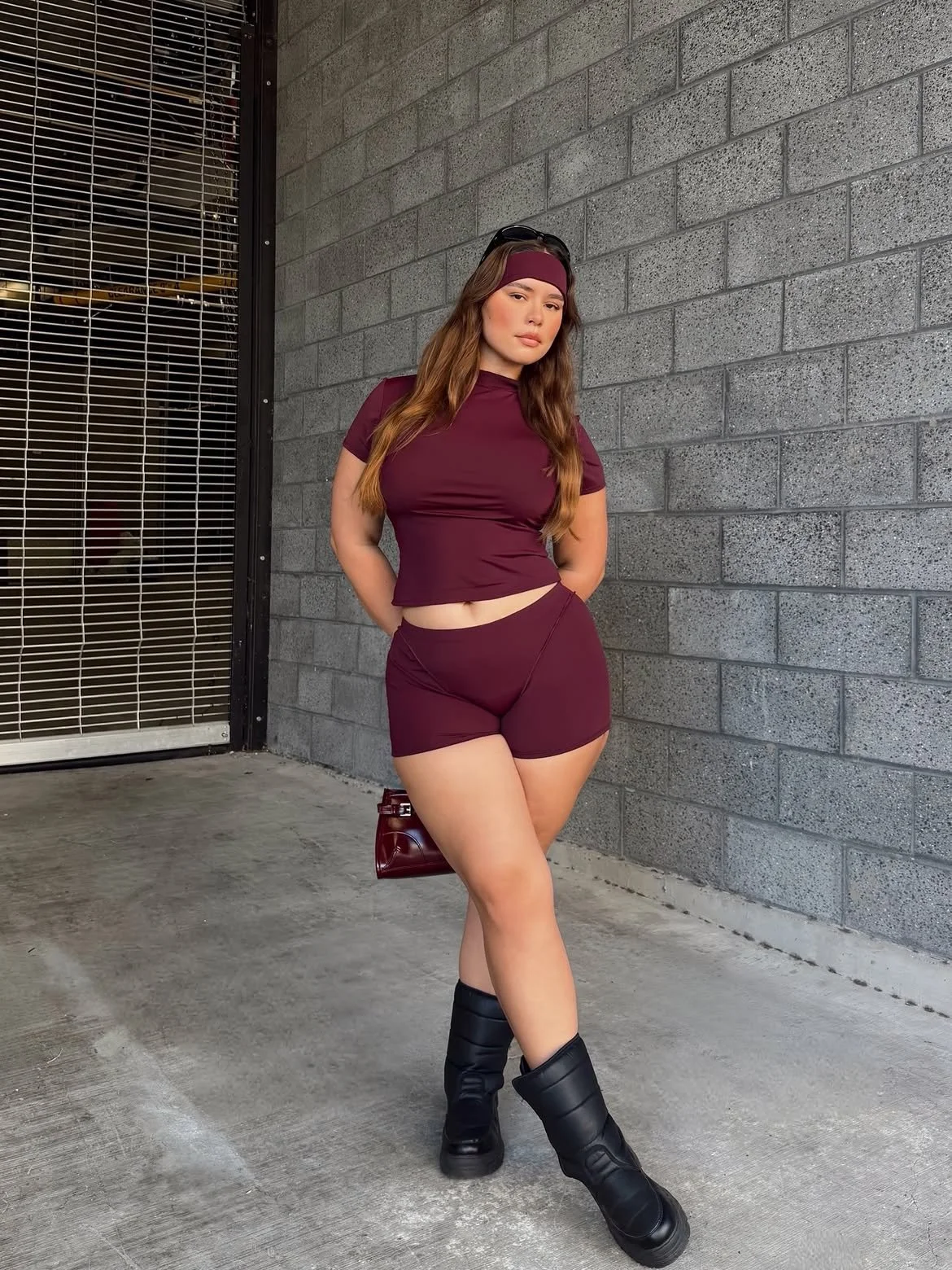 A young woman wearing a maroon athletic outfit, including a matching crop top, shorts, and a headband, standing against a gray brick wall with her hands behind her back, holding a small maroon purse, with black boots and sunglasses on her head.