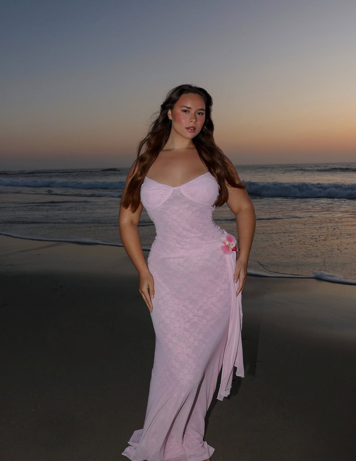 A woman in a pink dress standing on the beach at sunset with waves in the background.