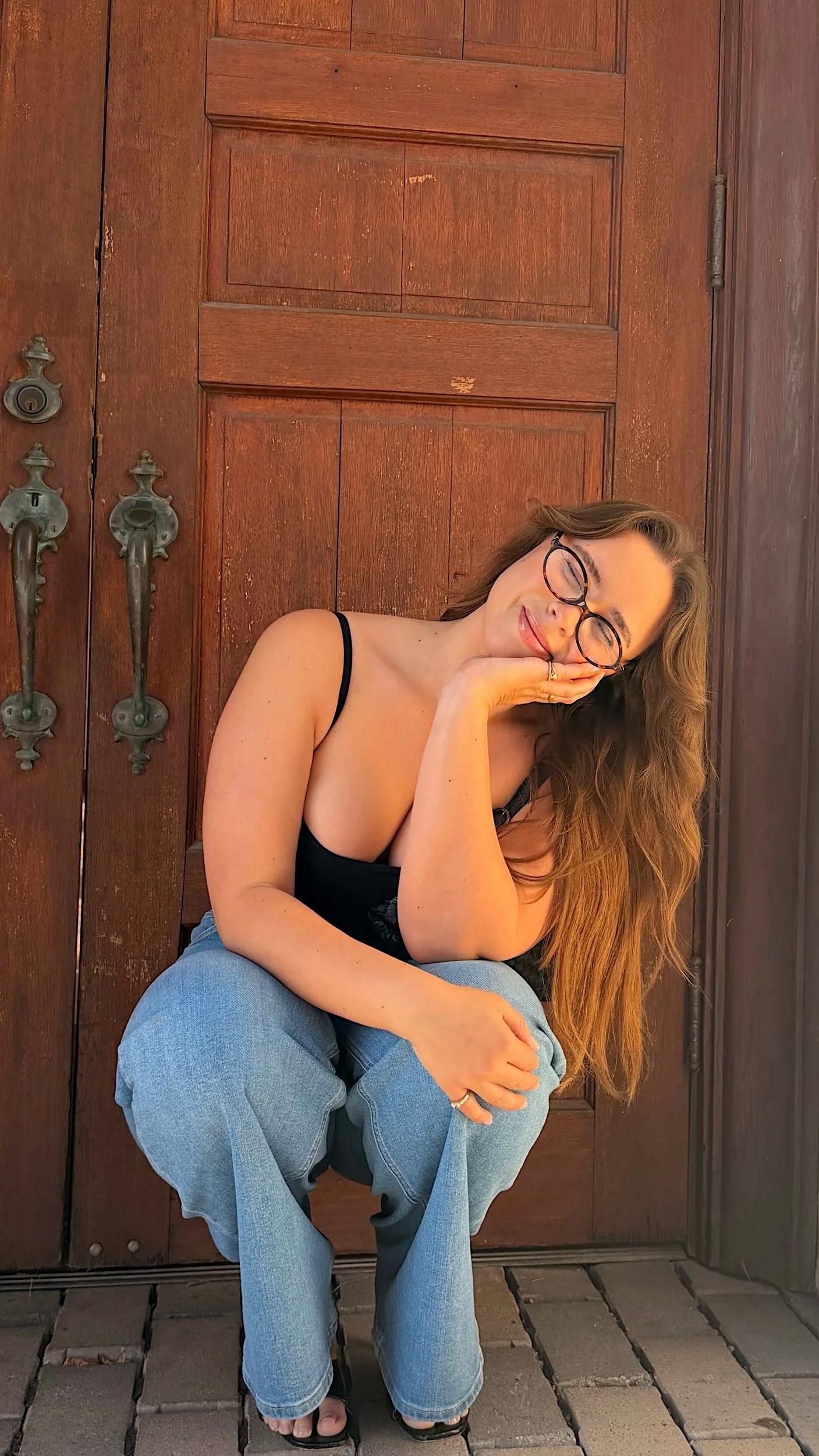 A woman with long, wavy hair and glasses sits in front of a wooden door, resting her head on her hand and smiling with her eyes closed. She is wearing a black spaghetti strap top and light blue jeans.