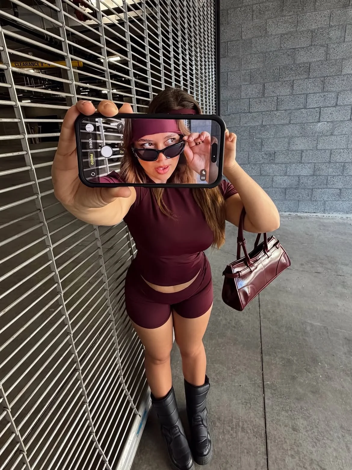 A woman taking a selfie with her phone, standing outdoors against a metal fence and brick wall. She is wearing sunglasses, a matching burgundy athletic outfit, combat boots, a headband, and carrying a burgundy purse.
