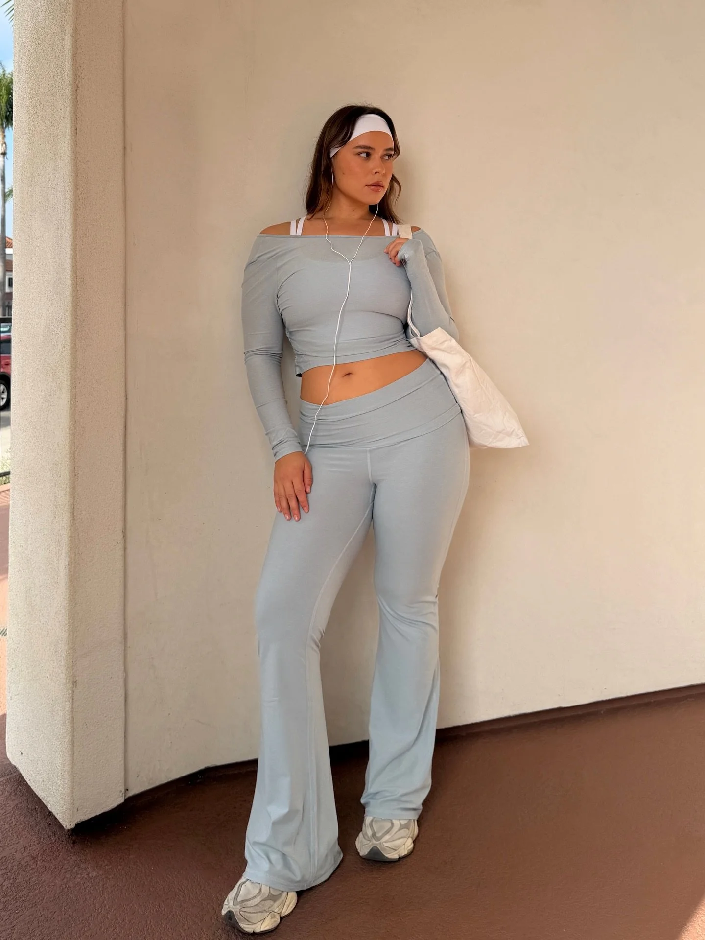A woman leaning against a beige wall, wearing a light grey off-the-shoulder crop top, matching grey sweatpants, white sneakers, a white headband, and headphones, with a white bag slung on her shoulder.