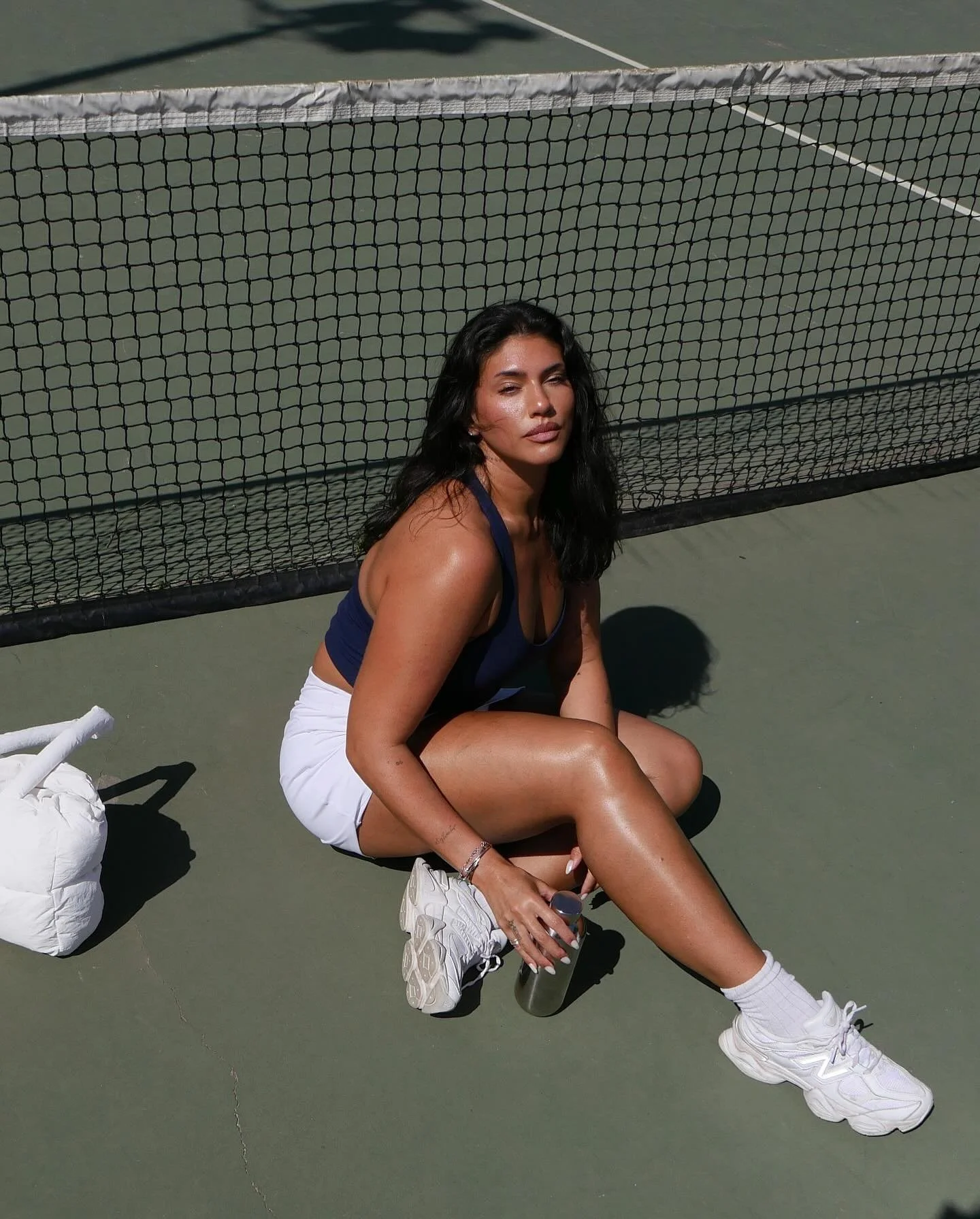 A woman with long black hair, wearing a navy blue athletic top and white shorts, sitting on a tennis court next to a net, holding a water bottle and a smartphone.