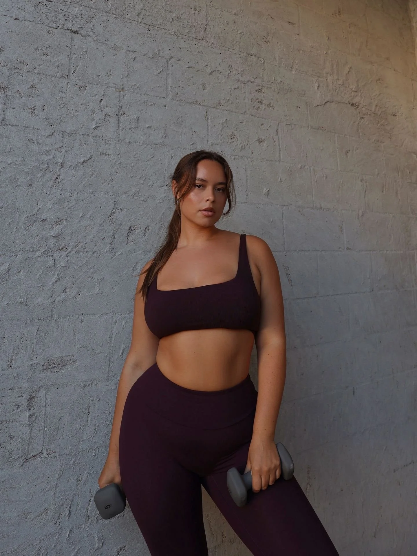 Young woman in workout clothes holding dumbbells against a brick wall at sunset