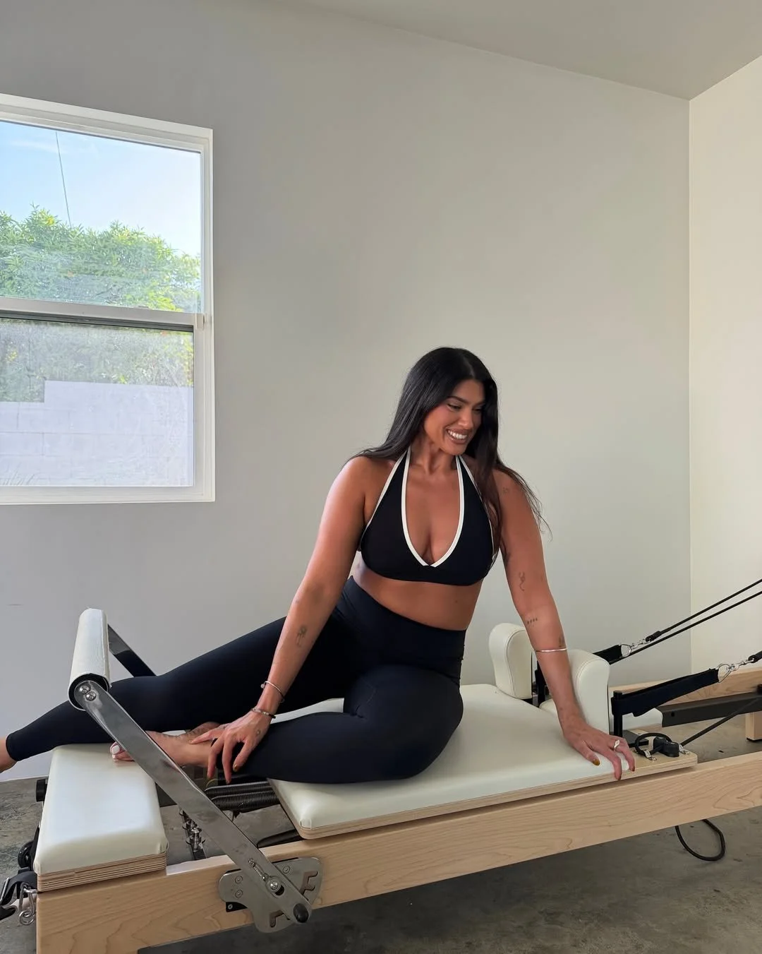 A woman in workout attire sitting on a Pilates reformer machine, smiling, in a bright room with a window showing greenery outside.