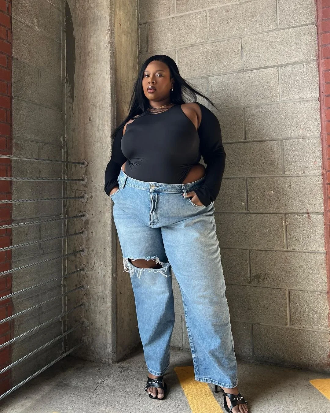 A woman with long black hair wearing a black top with cutouts and ripped jeans, standing against a concrete and brick wall in an urban setting.