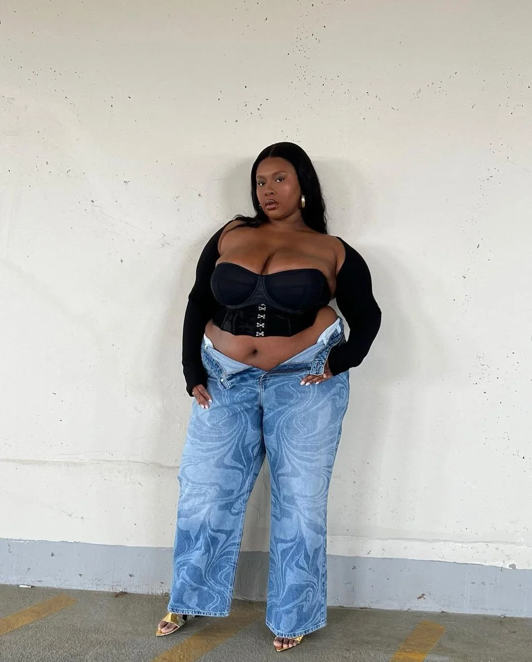 Woman posing against a plain white wall, wearing a black bustier top, long black cardigan, oversized blue patterned jeans, and heels.