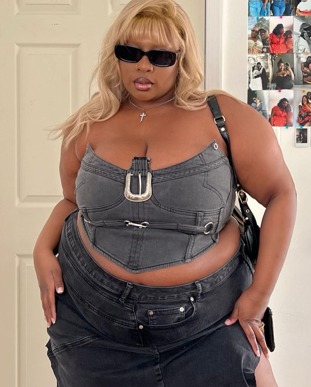 A woman with blond hair wearing black sunglasses, a cross necklace, a sleeveless denim top with belt details, and dark jeans, standing in front of a white door and a collage of photos on the wall.