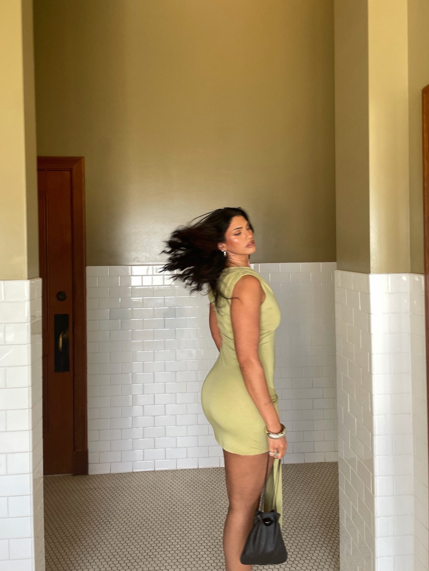 A woman with dark hair wearing a yellow dress standing in a tiled bathroom with her hair blowing, holding a black handbag.