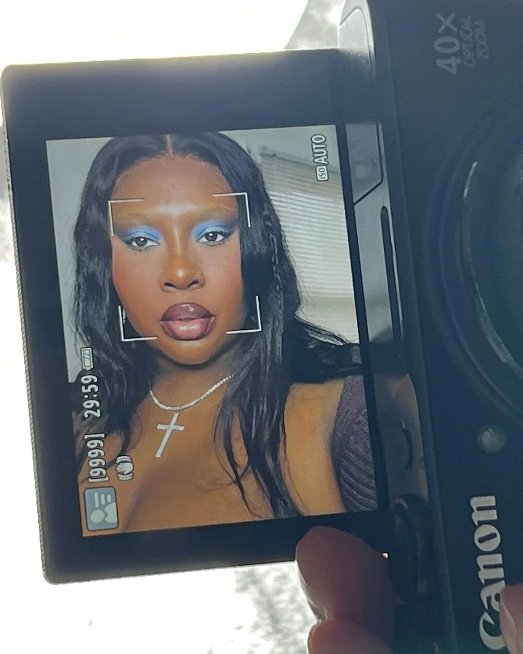 Digital camera screen displaying a woman with makeup, wearing a necklace with a cross, and posing for a portrait.