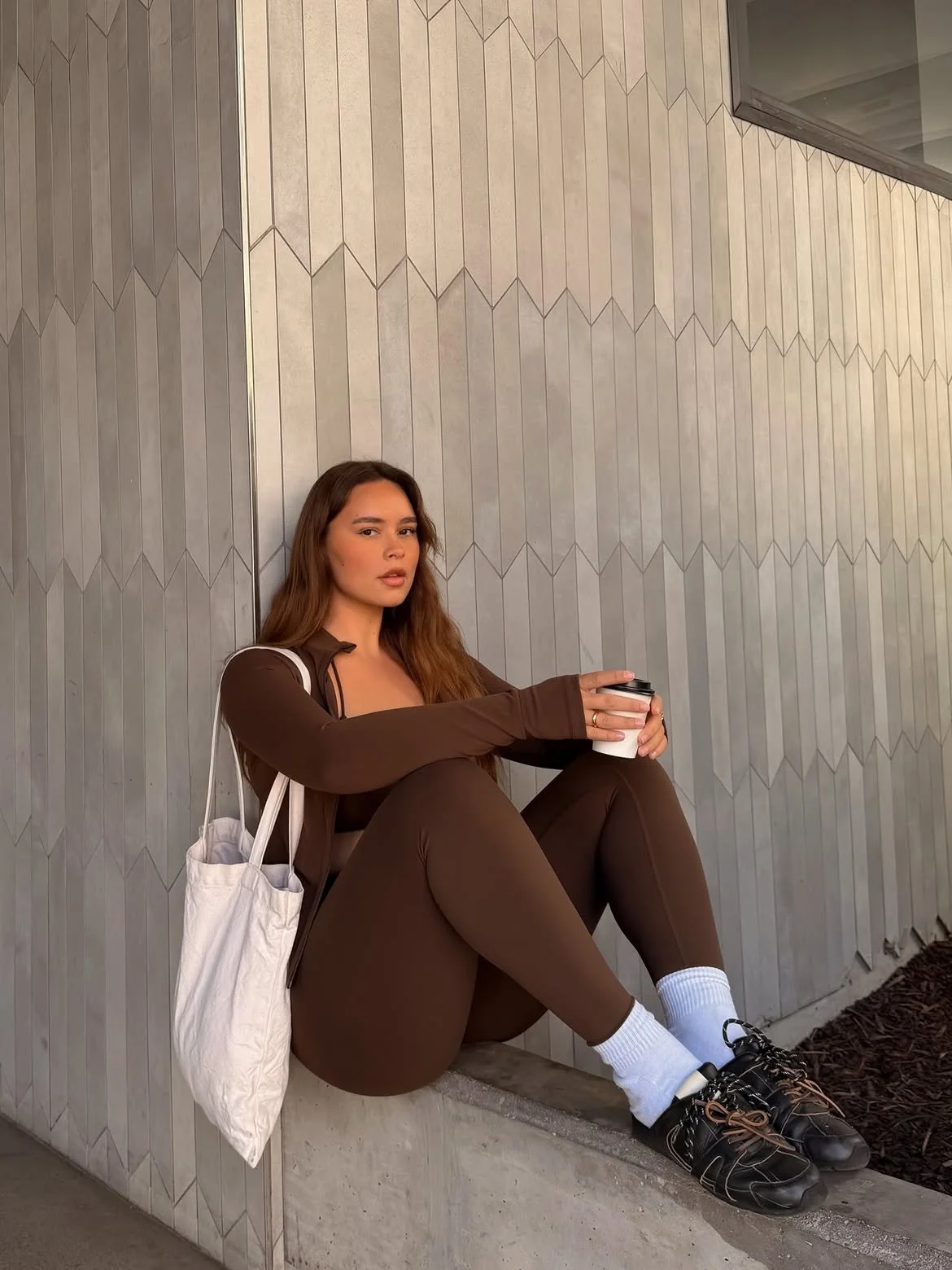 A woman sitting against a geometric patterned wall, holding a coffee cup, wearing a brown outfit, white socks, and black shoes, with a white tote bag on her shoulder.