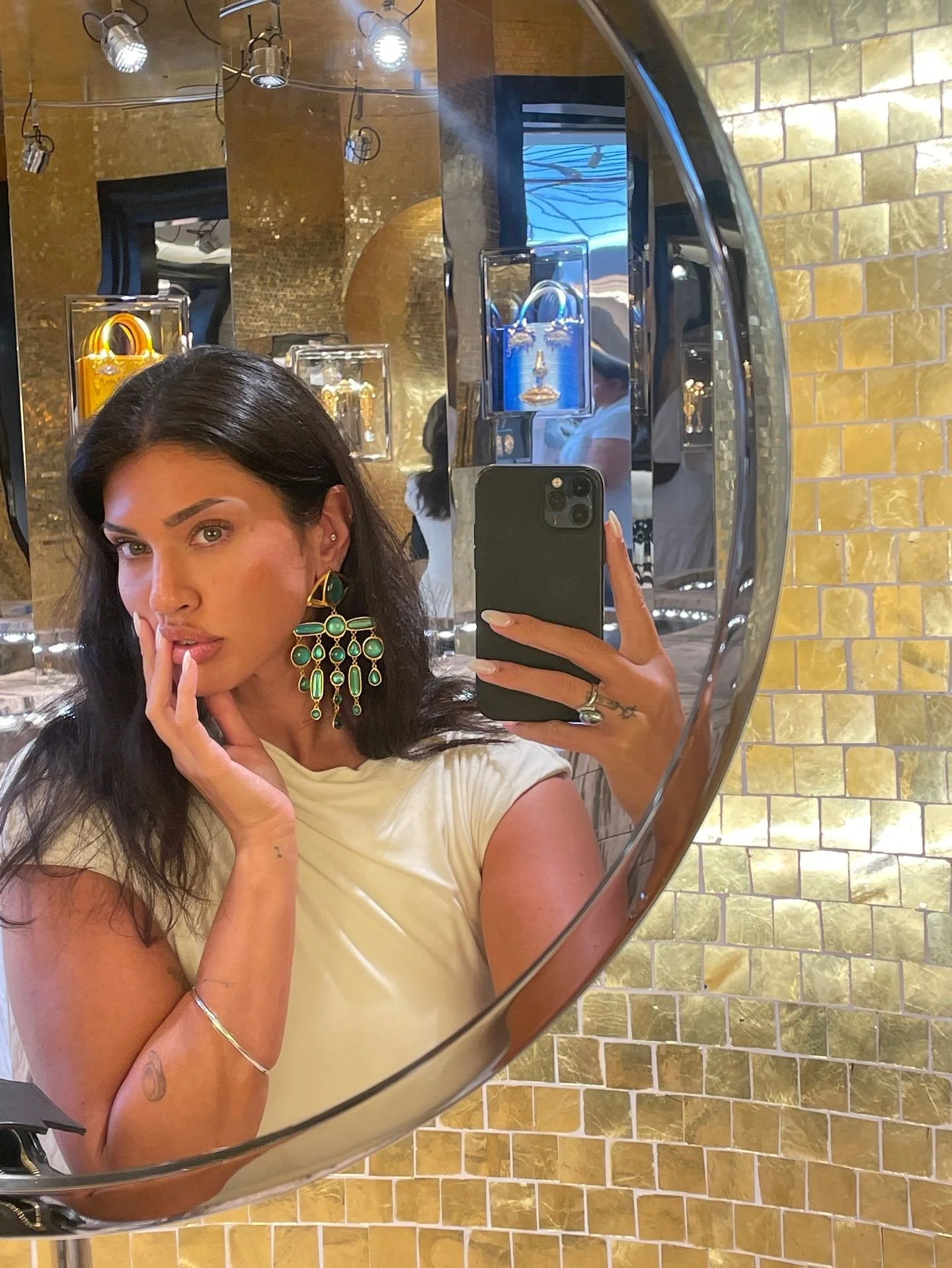A woman is taking a selfie in a mirror at a jewelry store, with display cases of handbags behind her. She is wearing large, green jewelry earrings, a beige dress, and has dark hair. The background features a yellow brick wall and lighting fixtures.